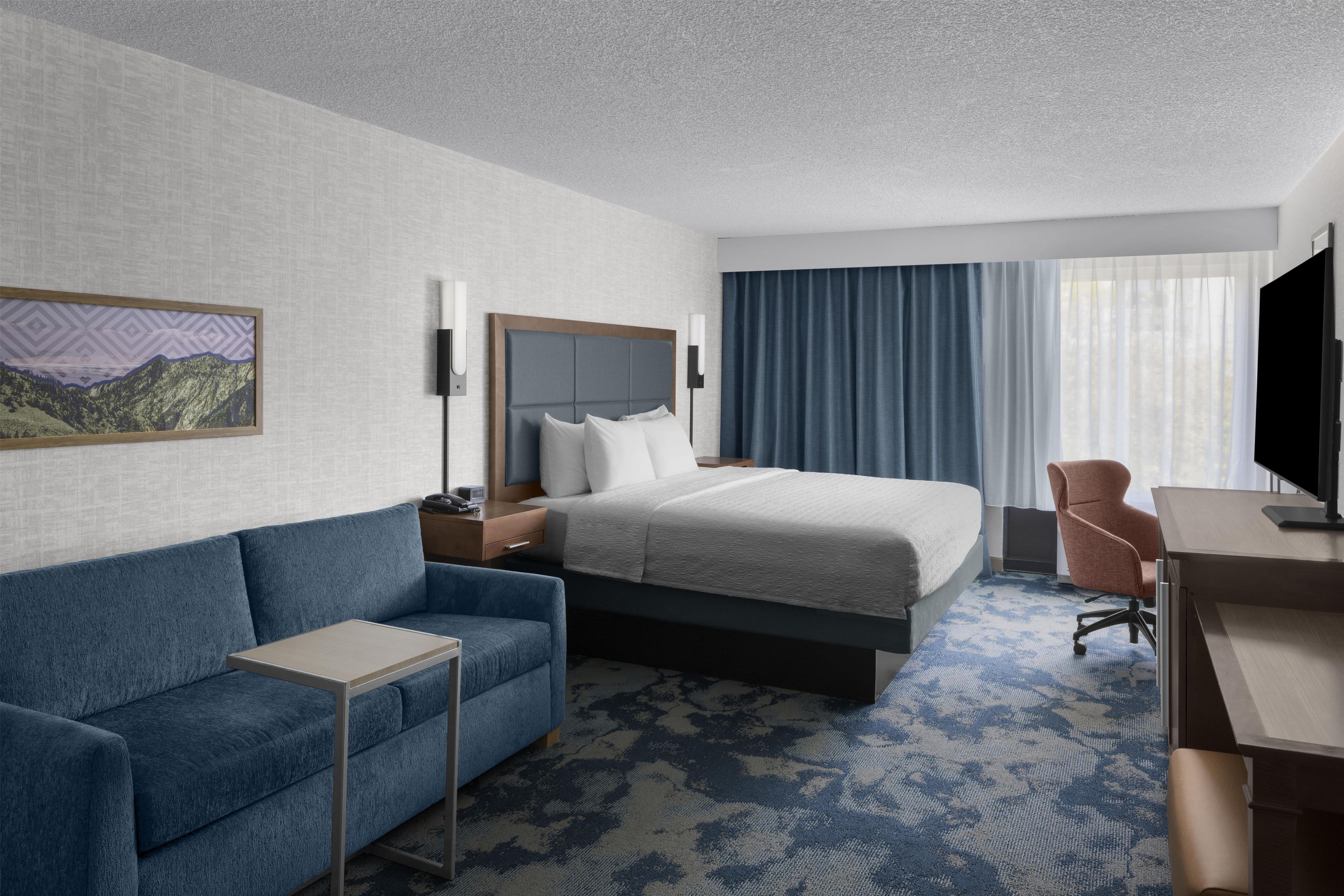 Quarto de hotel com cama, escrivaninha, cadeira, mesa pequena e televisão.