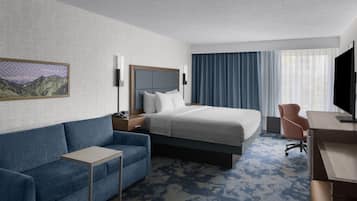 Quarto de hotel com cama, escrivaninha, cadeira, mesa pequena e televisão.
