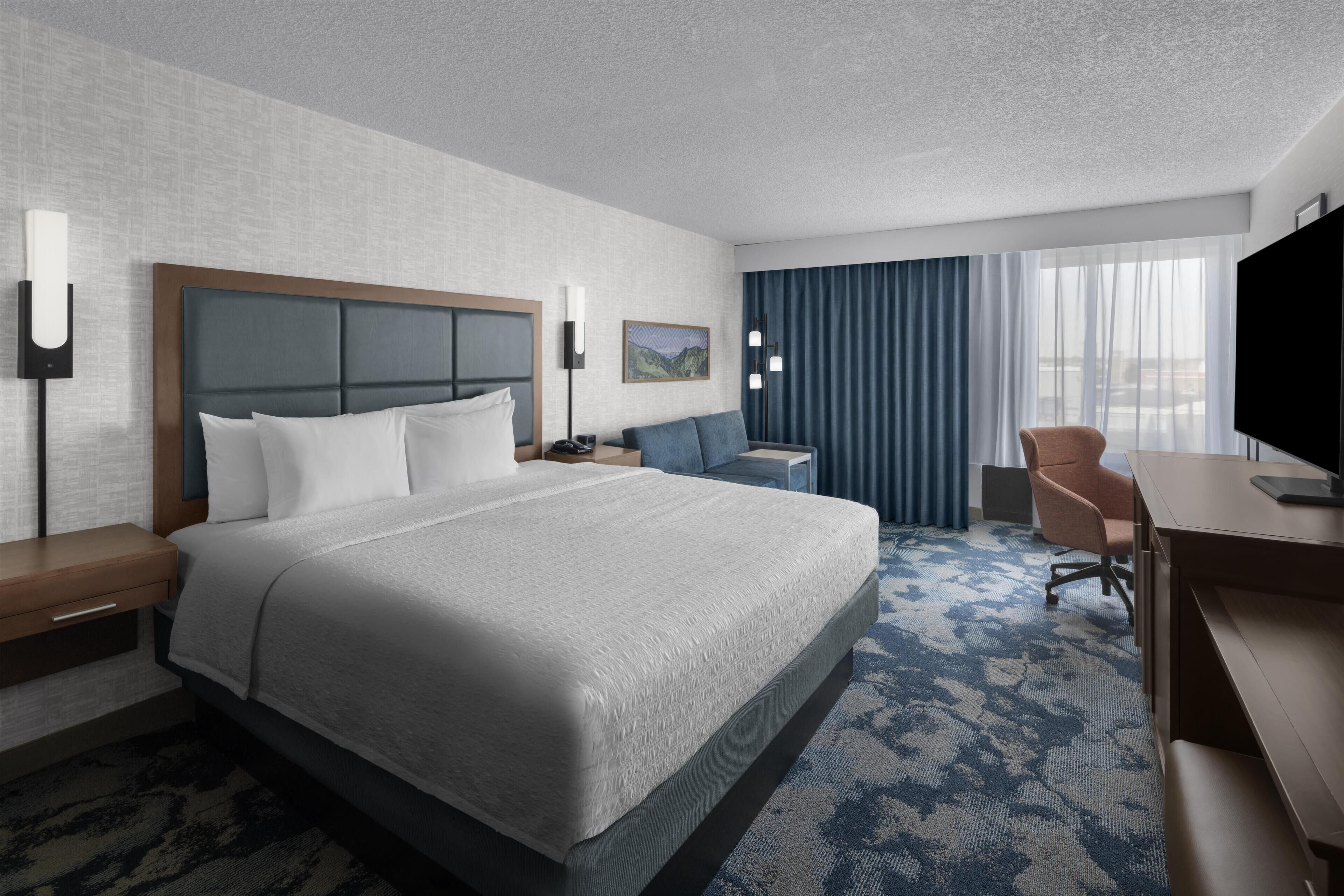 A hotel room with a large bed, a desk, a chair, a TV, and a window with curtains.