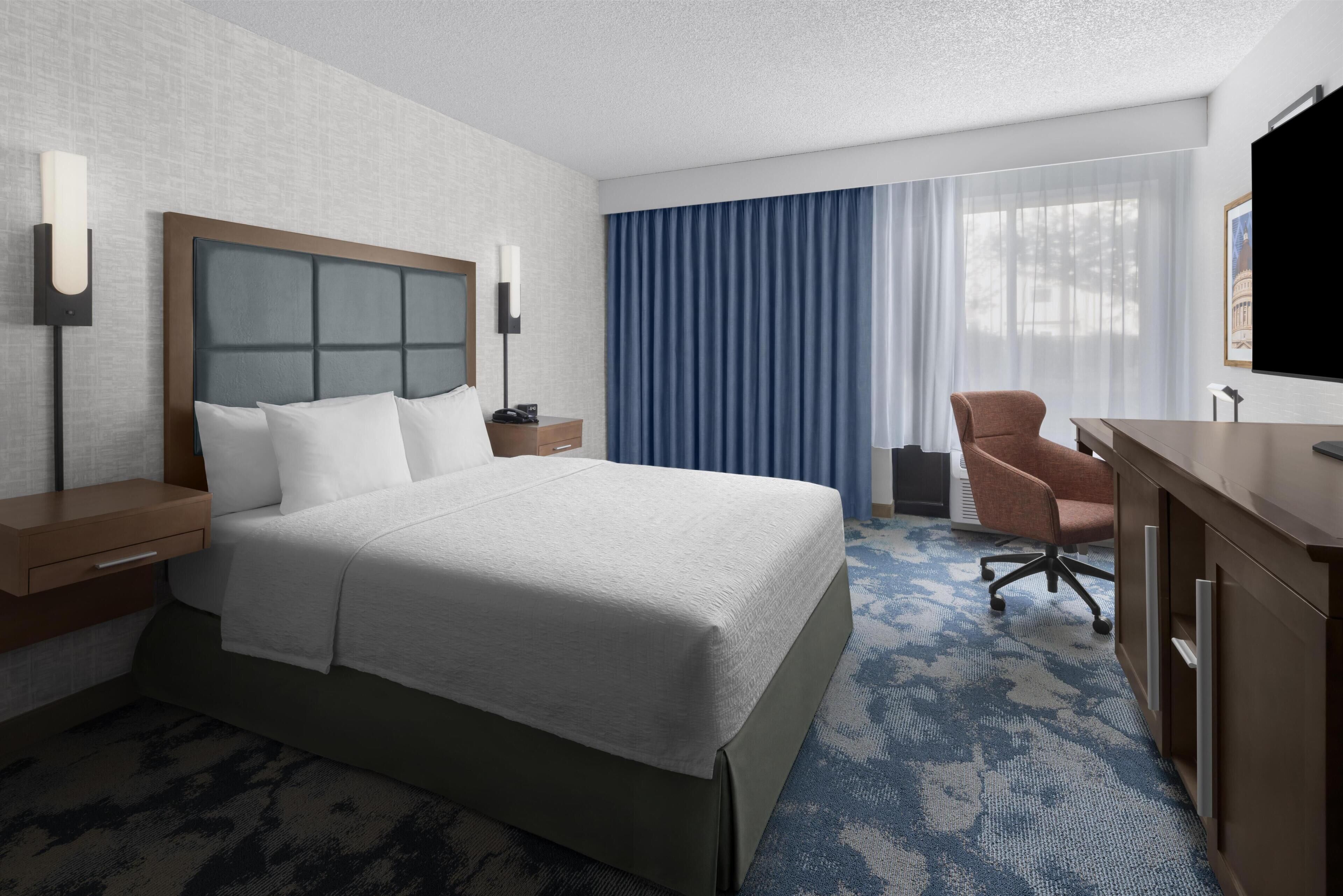 Habitación de hotel con una cama grande, un escritorio, una silla, un televisor y una ventana con cortinas.