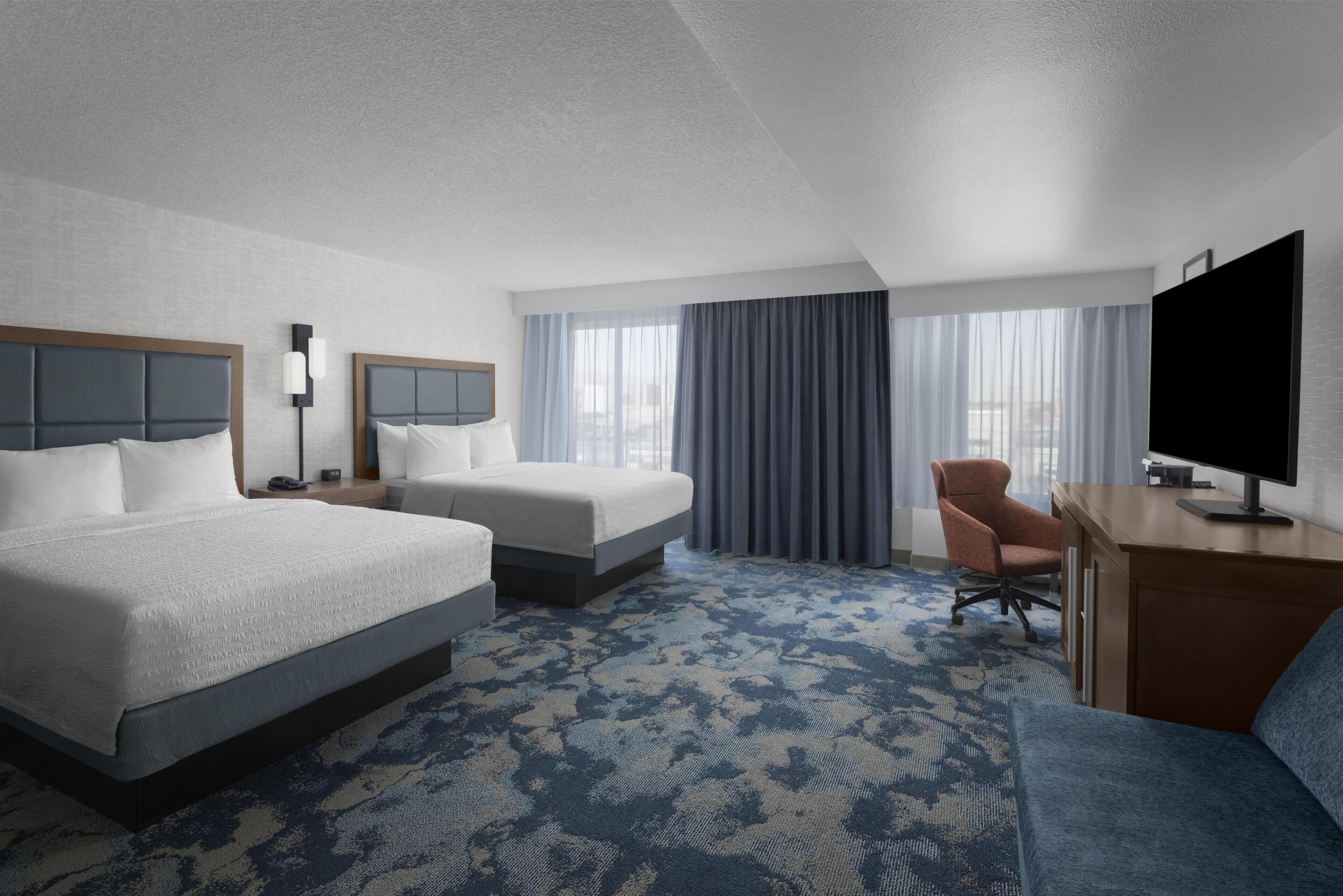 A hotel room with two beds, a desk, a chair, a TV, and a large window with curtains.