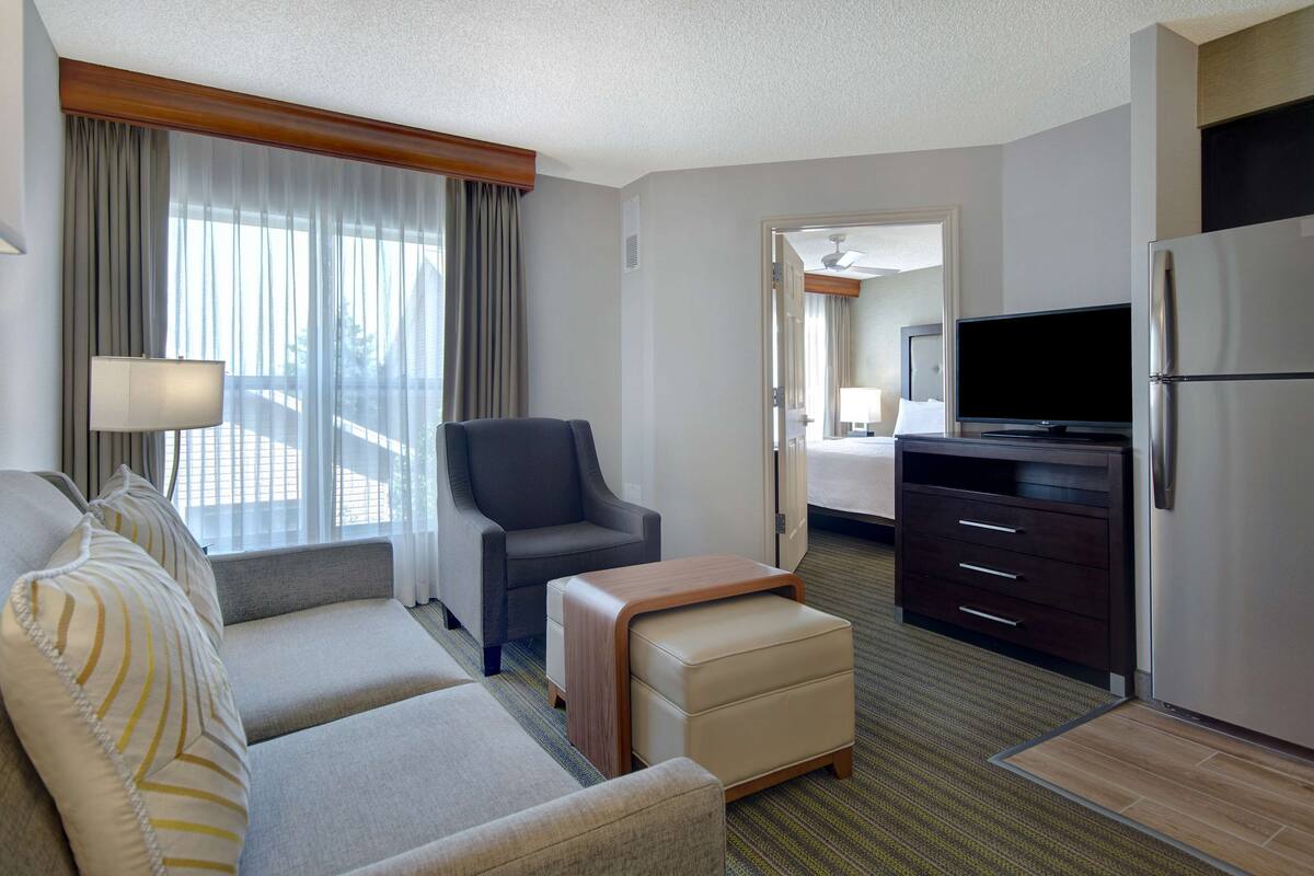 Ein Hotelzimmer mit Sofa, Sesseln, Hocker, Fernseher, Kühlschrank und Blick auf den Balkon durch leichte Vorhänge.