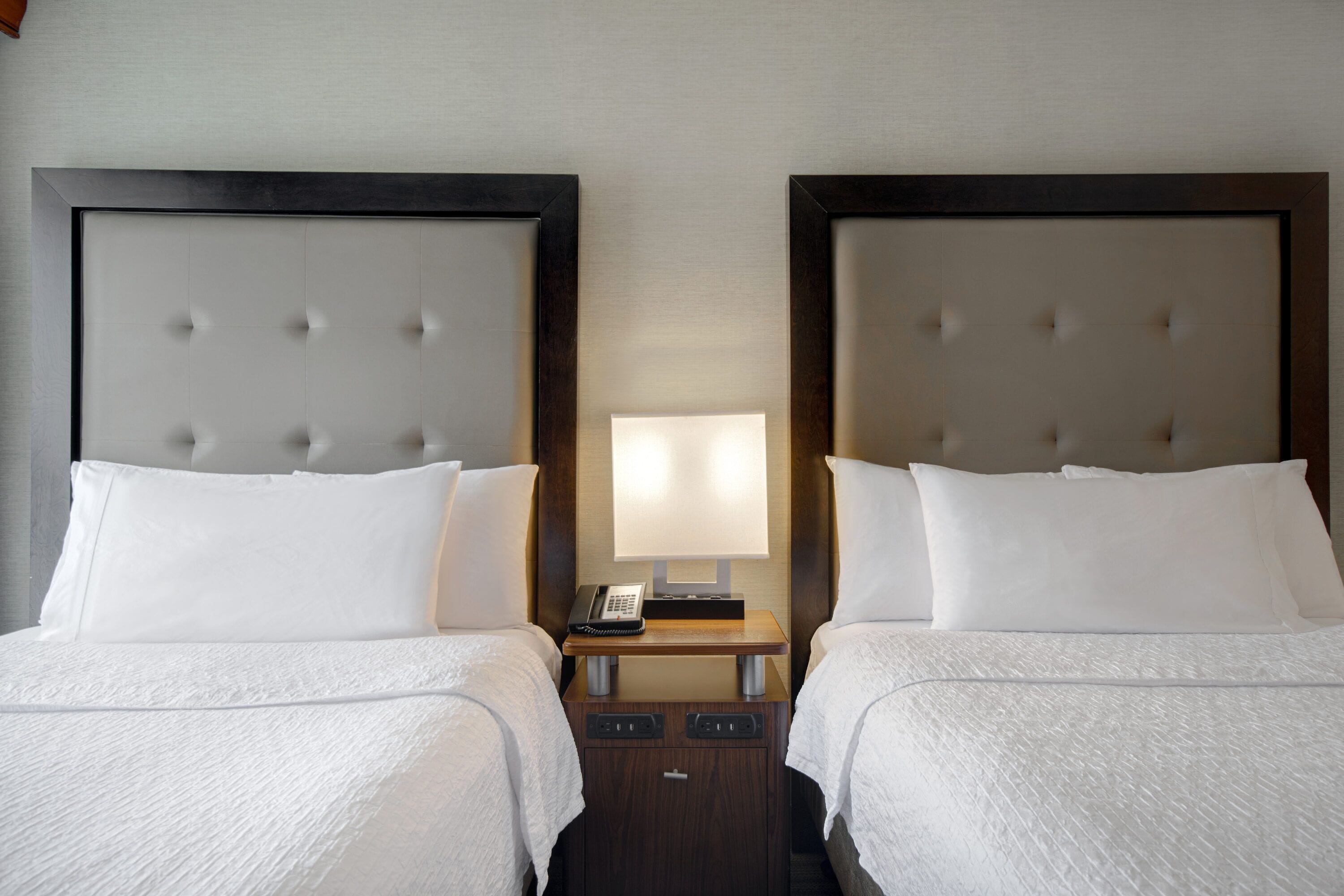 A hotel room with two beds, a bedside table with a lamp, and a telephone.