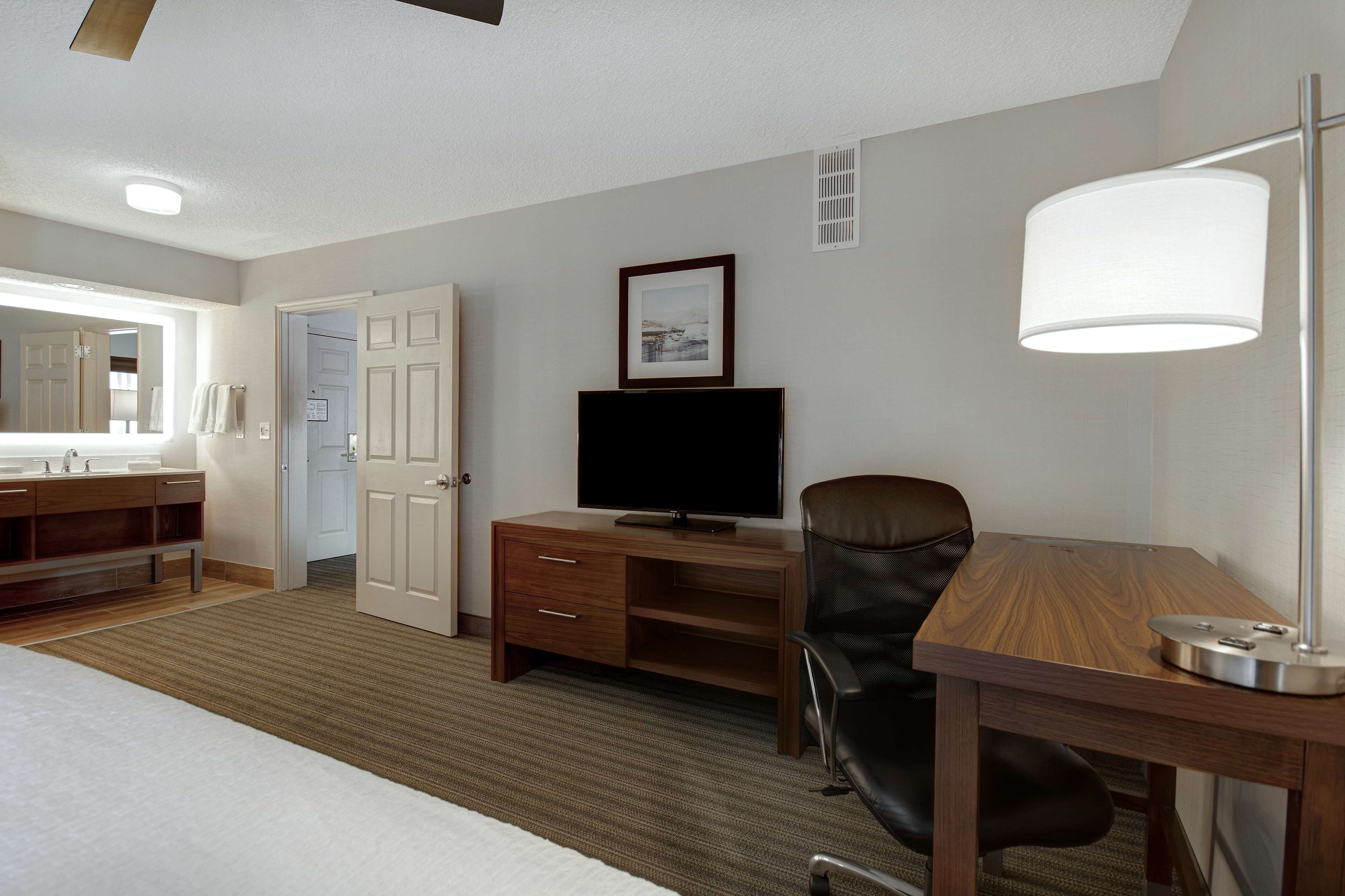 A hotel room with a bed, a desk with a lamp, a television, a chair, a wooden dresser, a bathroom with a sink and mirror, and a door leading to another room.