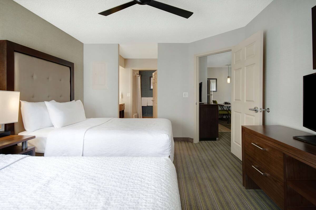 Ein Hotelzimmer mit zwei Betten, einem Deckenventilator, einer Holzkommode und einem Fernseher.