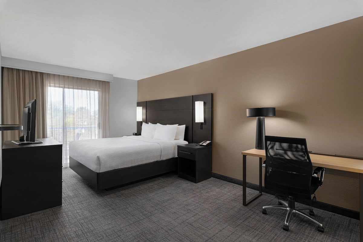 Ein Hotelzimmer mit Bett, Nachttisch, Schreibtisch, Stuhl und einem großen Fenster mit Vorhängen.