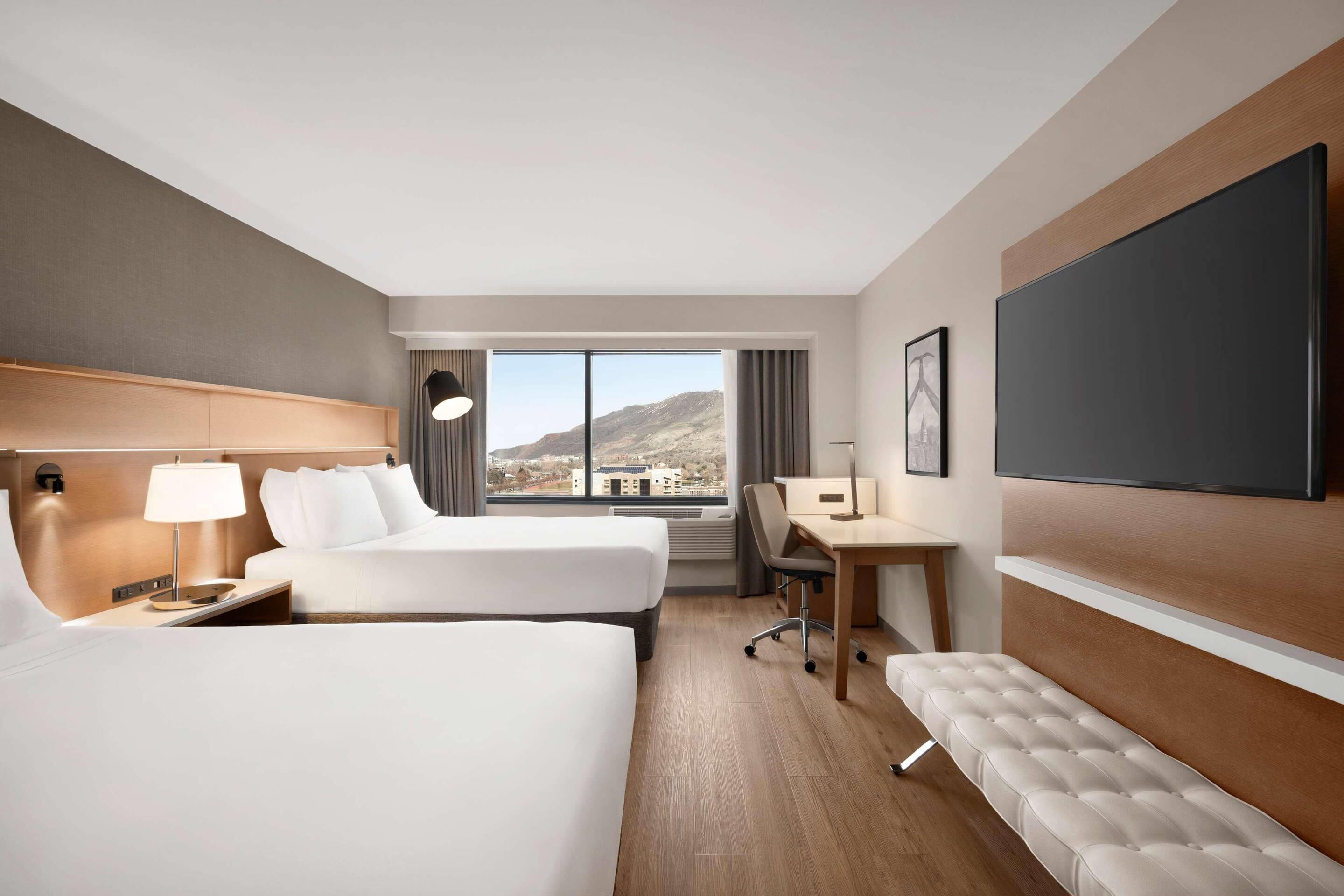 Quarto de hotel com duas camas, uma escrivaninha, uma cadeira, um banco, uma TV e uma janela grande com vista para as montanhas.