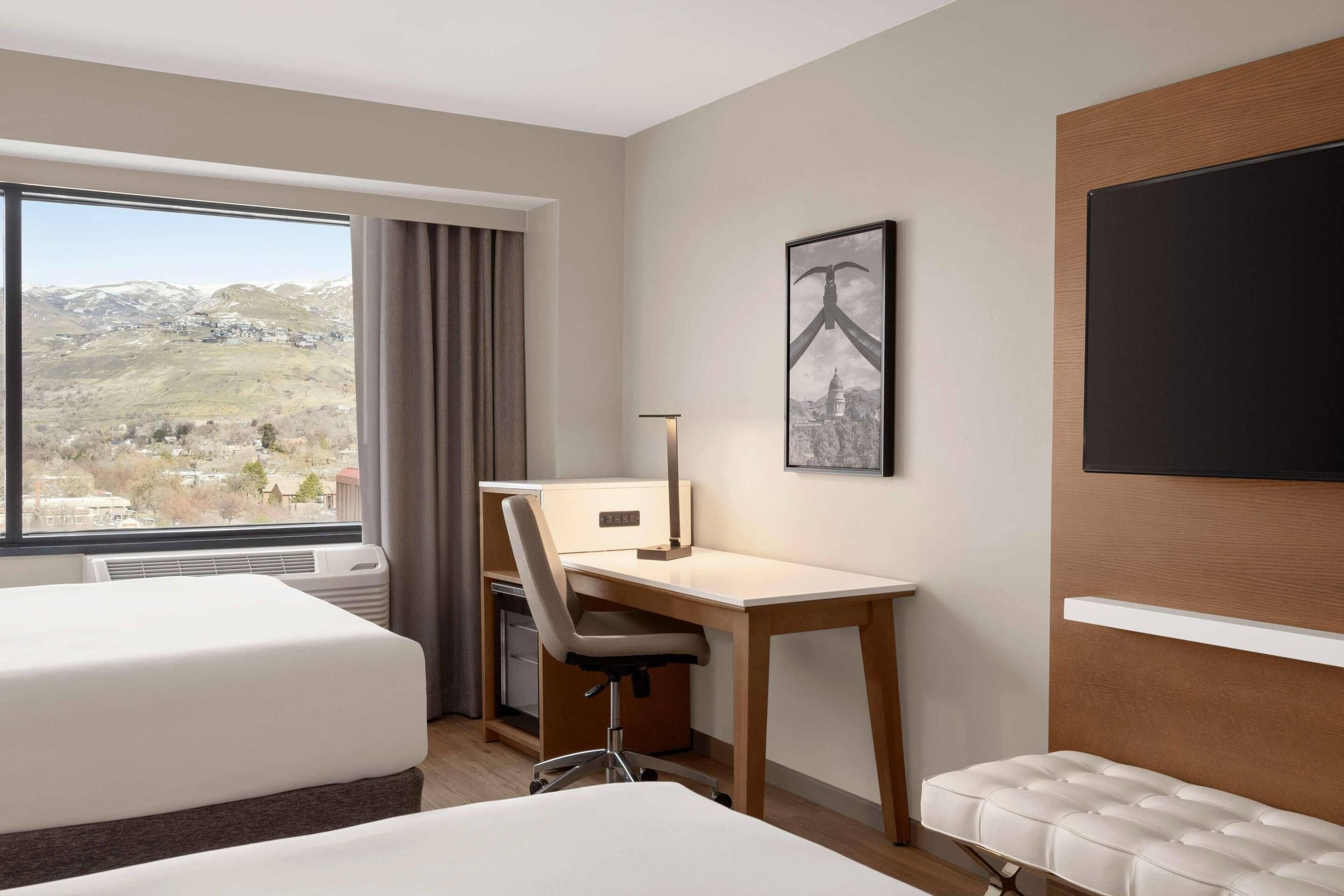 A hotel room with two beds, a desk, a chair, a TV, and a large window with a view of a town and mountains.