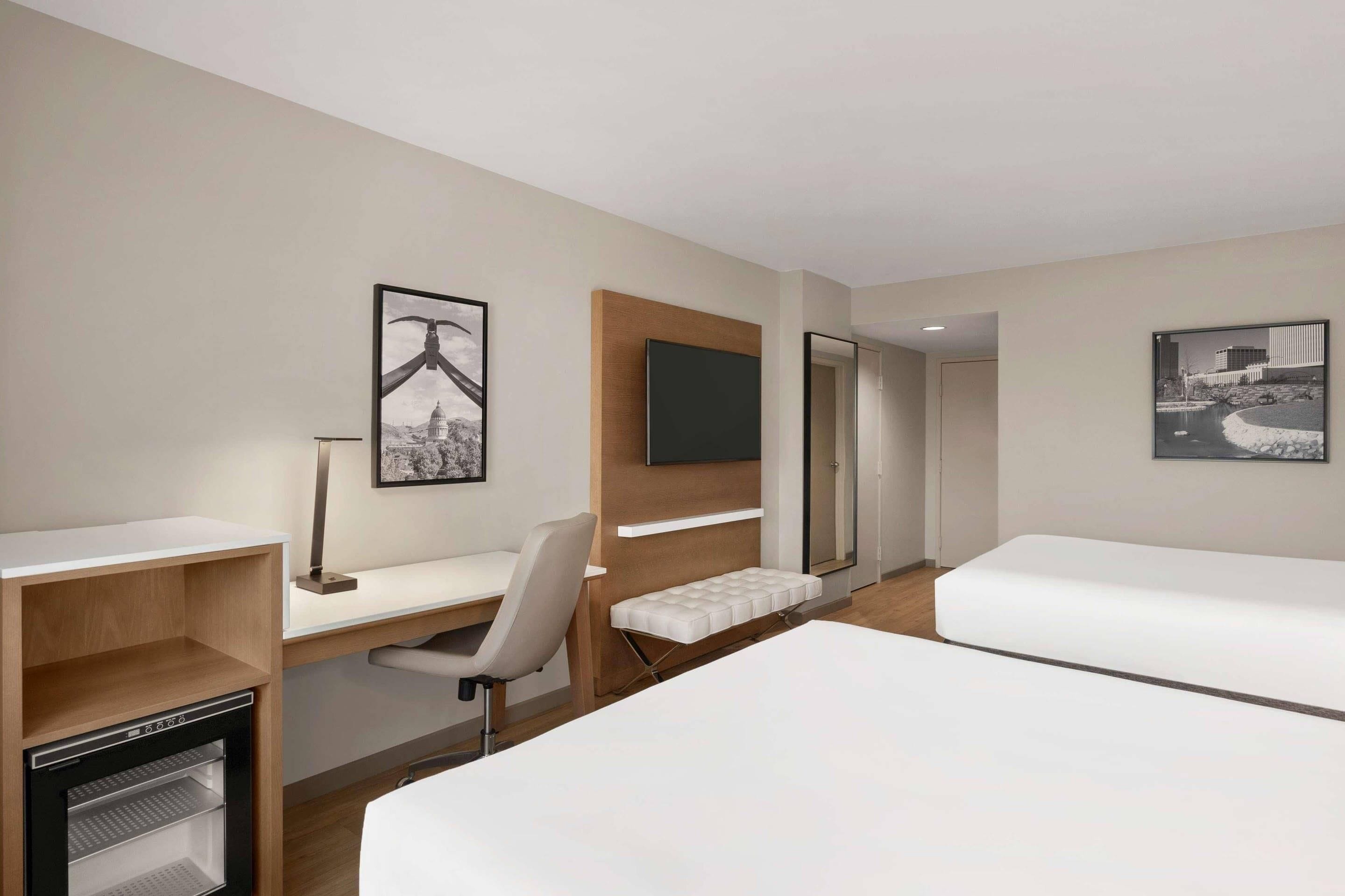 A hotel room with a bed, a desk, a chair, a small refrigerator, and two framed pictures on the wall.