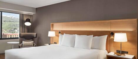 Habitación de hotel con una cama grande, dos mesitas de noche con lámparas, una silla y vistas a la montaña a través de la ventana.