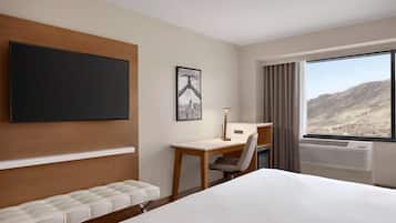 Habitación de hotel con una cama grande, un escritorio, una silla, un televisor de pantalla plana en la pared, un cuadro enmarcado y una ventana con vistas a la montaña.