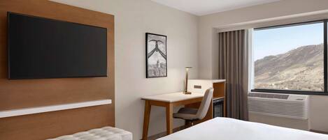 Habitación de hotel con una cama grande, un escritorio, una silla, un televisor de pantalla plana en la pared, un cuadro enmarcado y una ventana con vistas a la montaña.