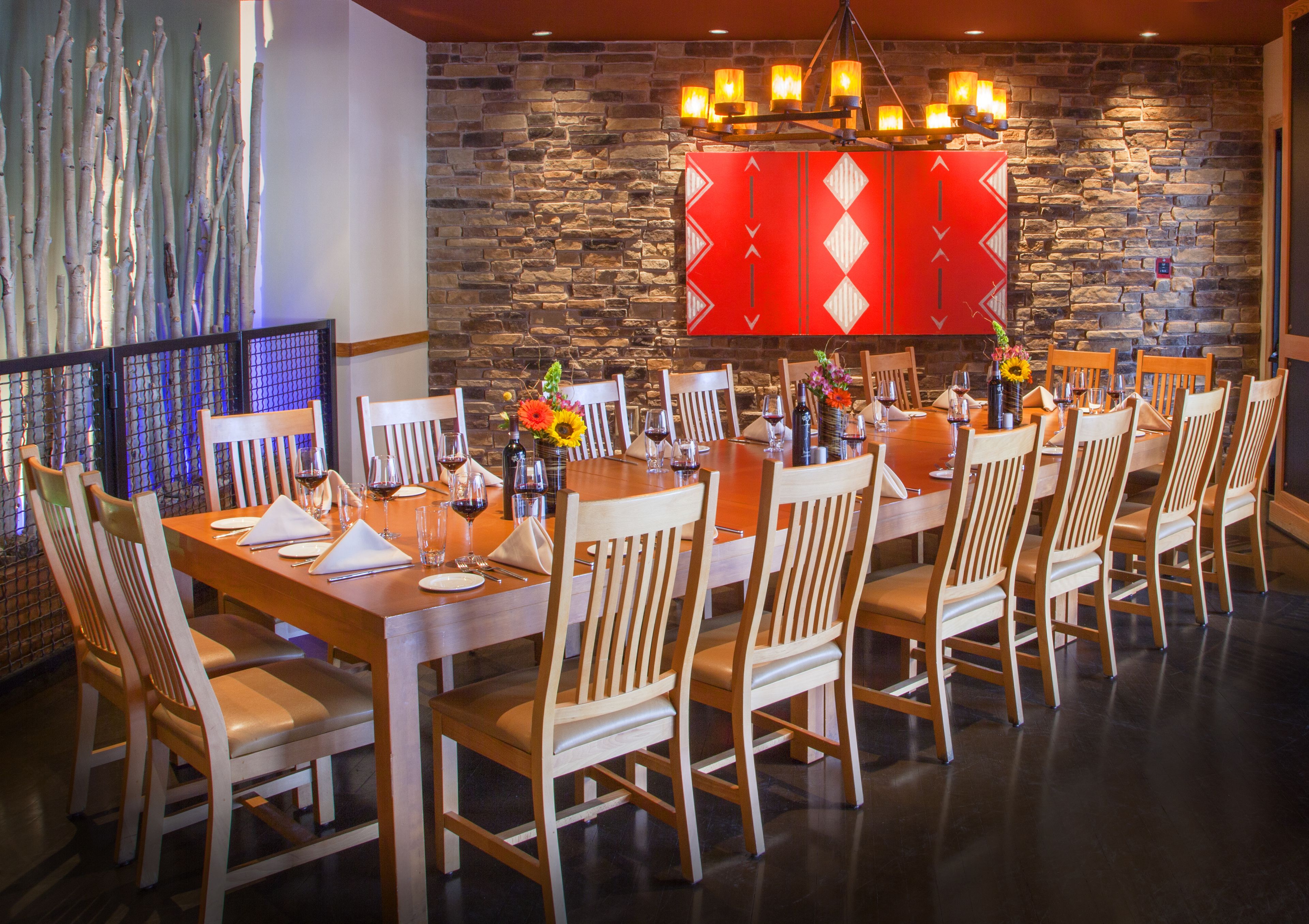 A dining area with a stone wall, wooden chairs, a long table set for a meal, and a decorative red panel above the table.