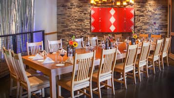 A dining area with a stone wall, wooden chairs, a long table set for a meal, and a decorative red panel above the table.