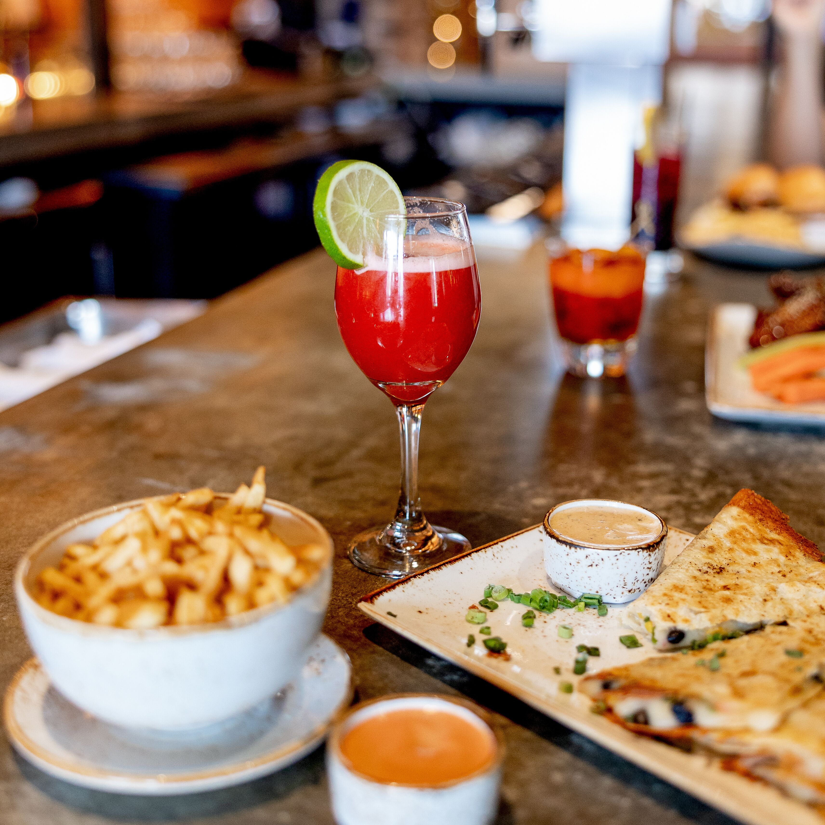Een barsetting met een glas rode cocktail, gegarneerd met een partje citroen, een schaal friet, een klein schaaltje saus, en een plak pizza op een bord.