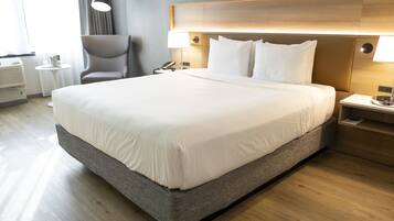 A neatly made bed with a wooden headboard, a bedside table with a lamp, and a chair in a modern hotel room.