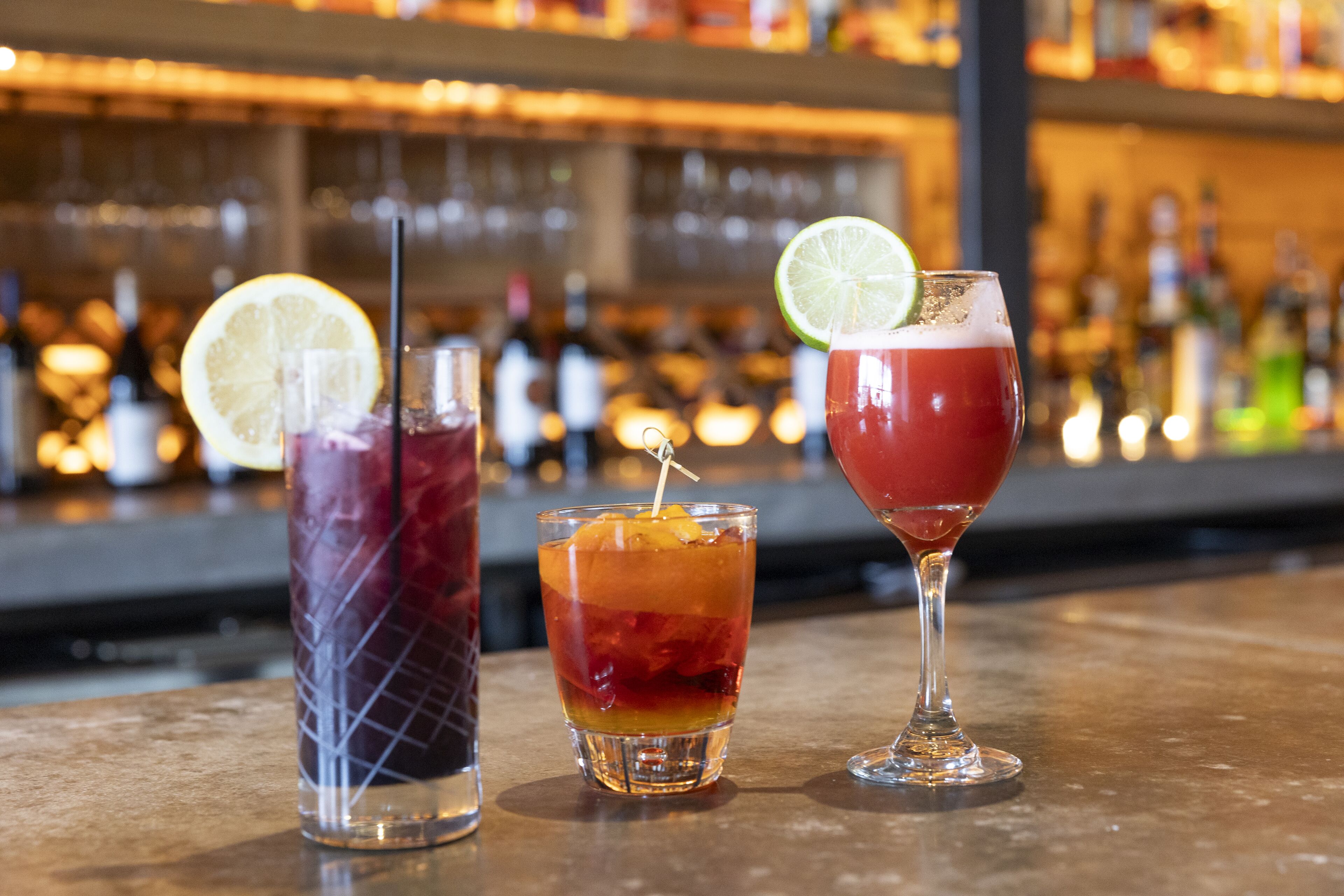Três coquetéis em um balcão de bar, com garrafas ao fundo, desfocadas.