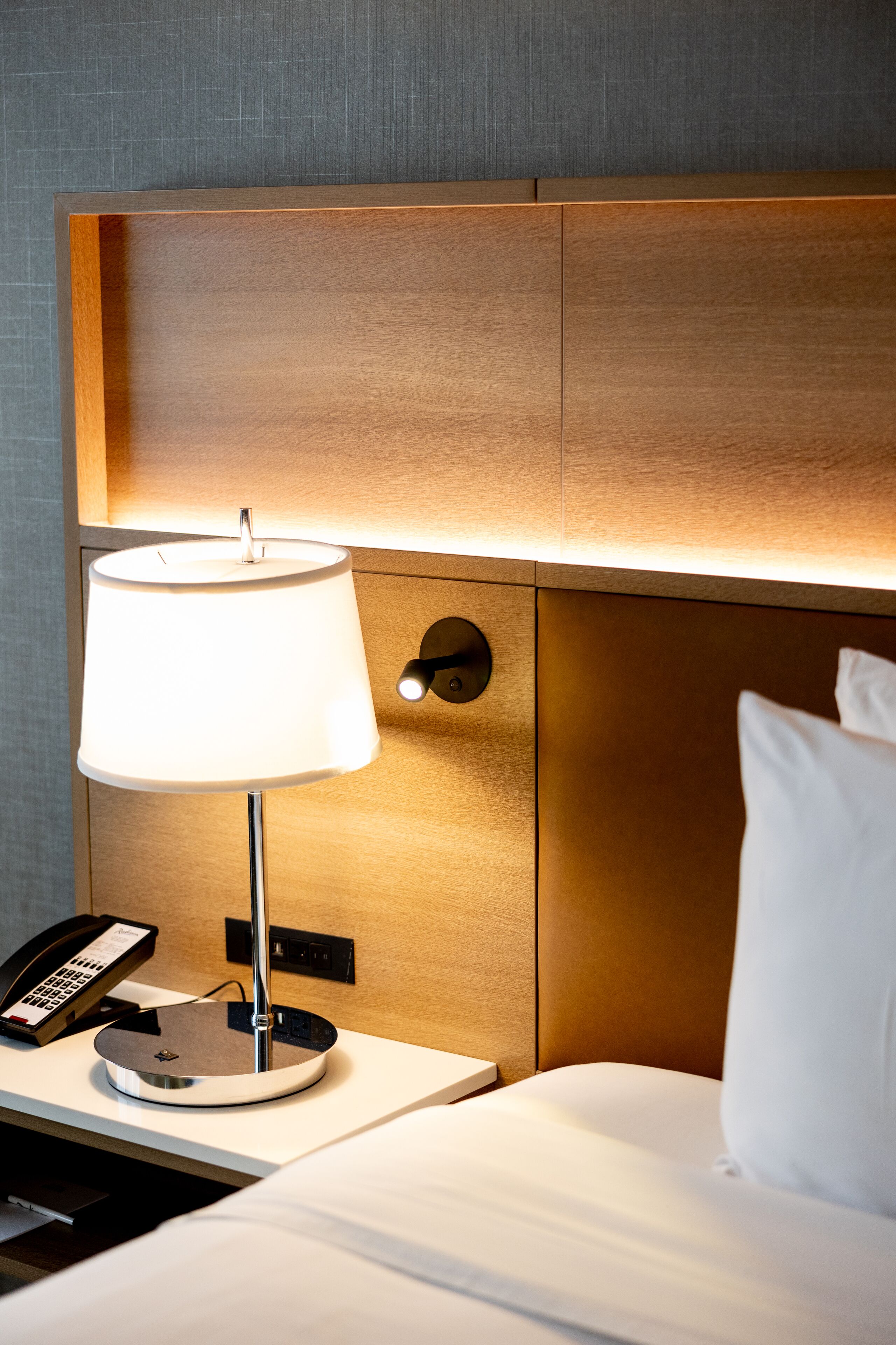 Una cama bien hecha con sábanas blancas, una lámpara de noche y un teléfono en una mesita de noche.