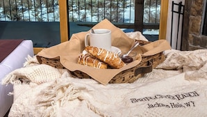 A cozy room with a view of snow-capped mountains, a wicker basket with pastries and a cup of coffee, and a blanket with "Sprinting Creek Ranch Jackson Hole, WY" embroidered on it.
