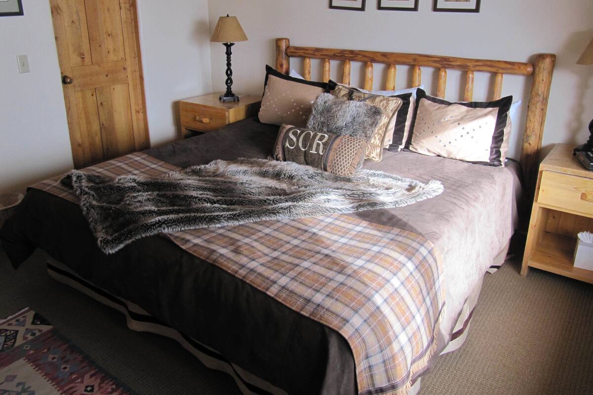A neatly made bed with a plaid blanket, a decorative pillow with 'SCR' embroidered, wooden headboard, and bedside tables with lamps.