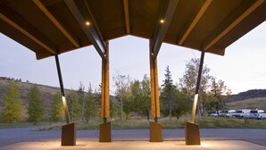 A covered outdoor area with a wooden roof and metal beams, surrounded by trees and parked vehicles.