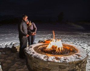 Two people standing by a stone fire pit with a burning fire in a snowy outdoor setting.