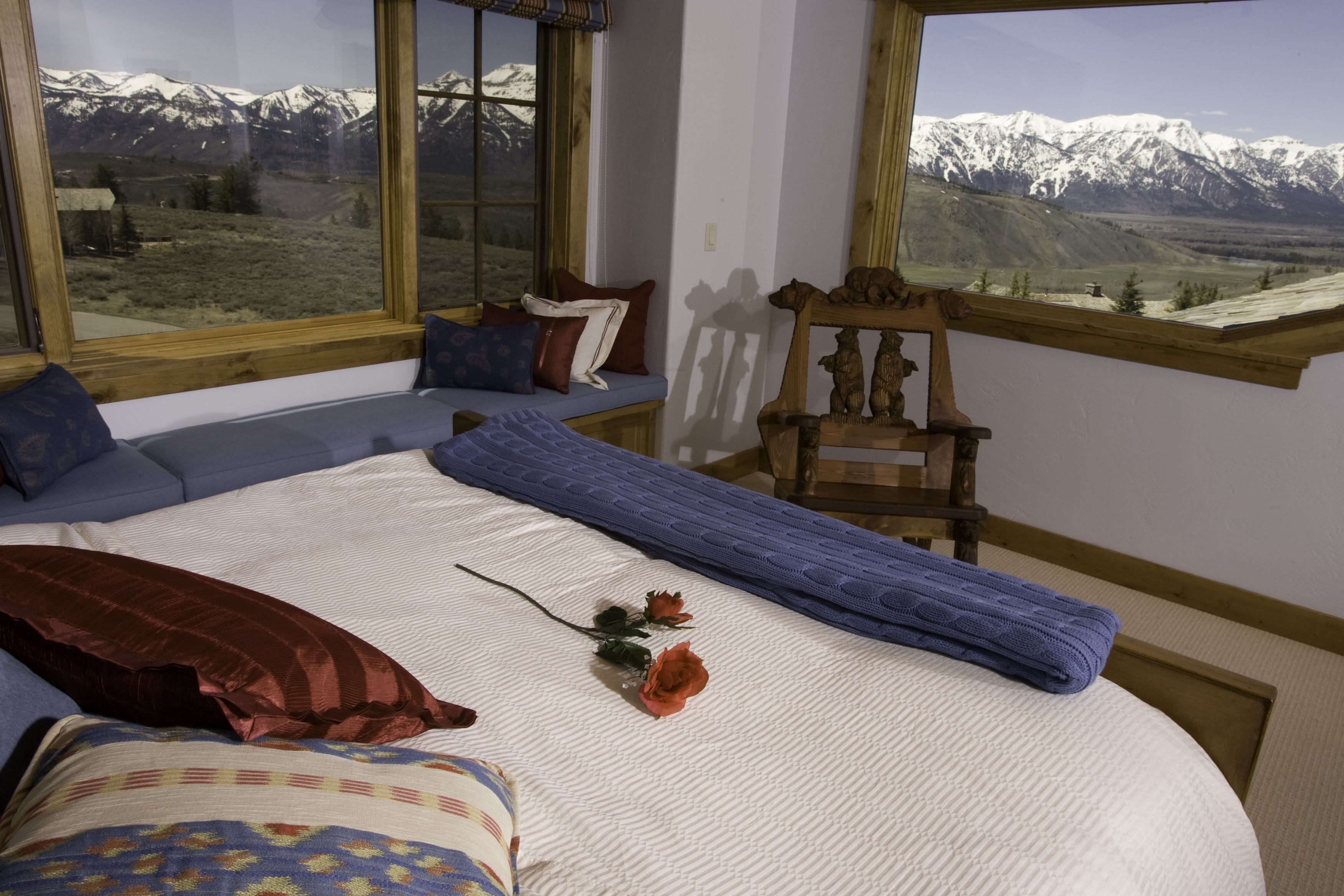 Un dormitorio con una cama grande, una silla de madera, un banco y vistas a montañas nevadas a través de amplias ventanas.
