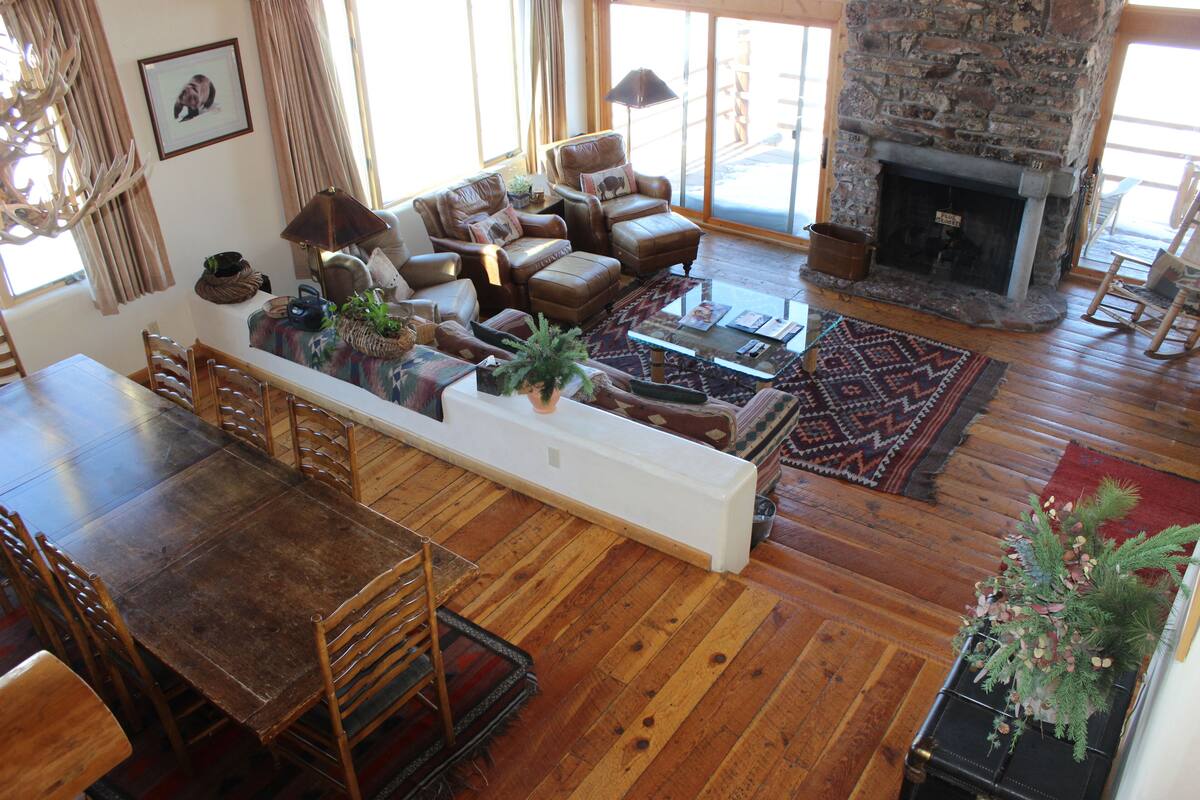 A spacious living room with a stone fireplace, wooden flooring, and a dining area with a long table and chairs.