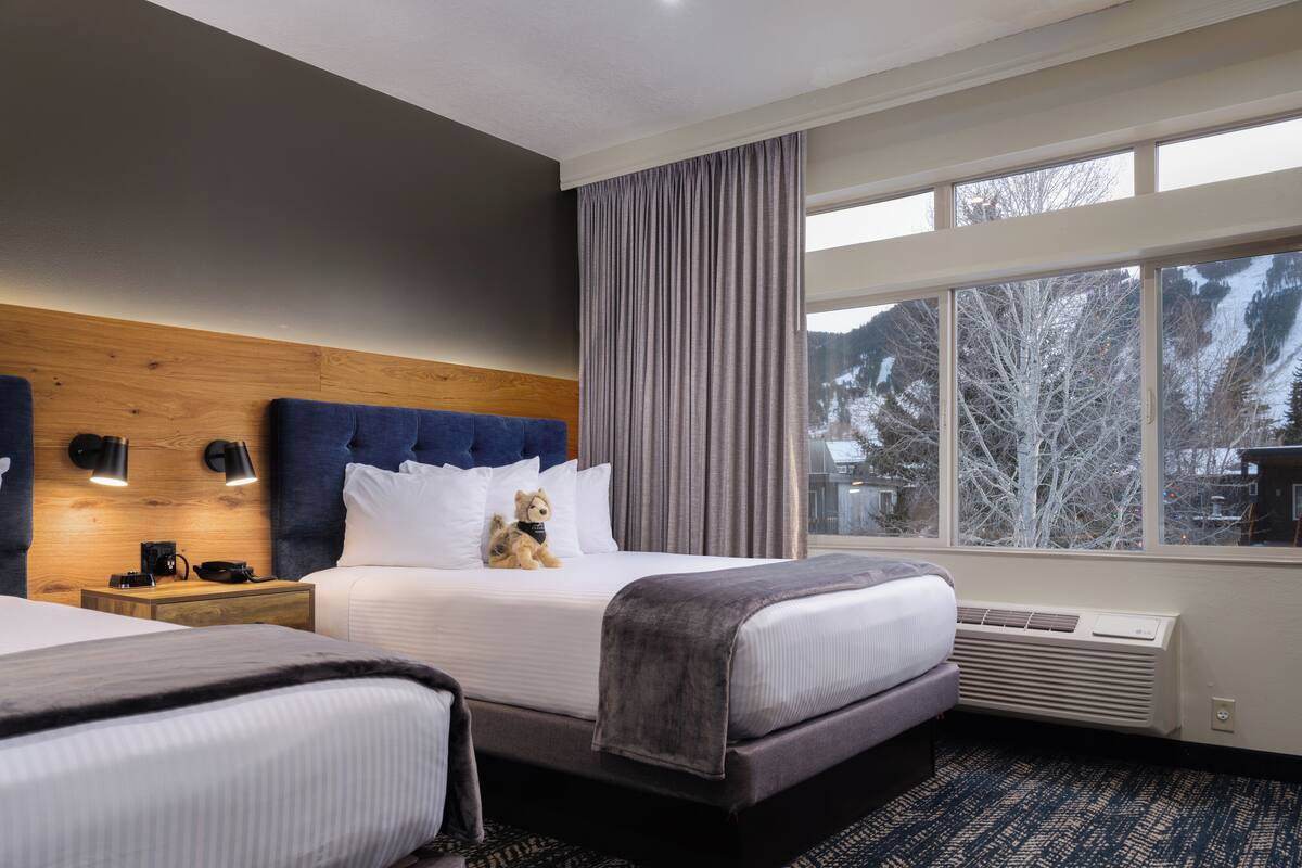 A hotel room with a large bed, a smaller bed, a wooden headboard, a nightstand, a lamp, a window with a view of trees, and a patterned carpet.