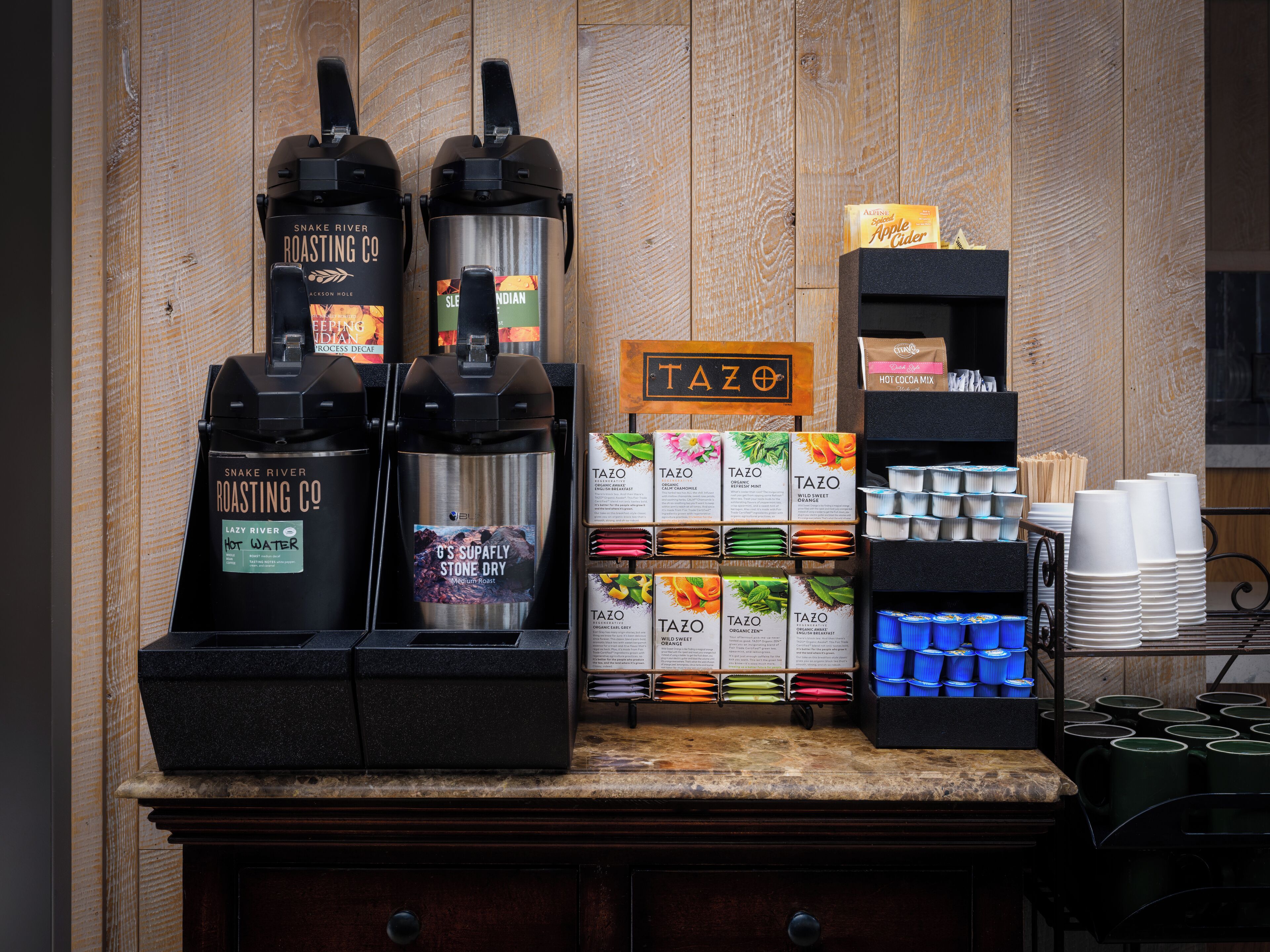 A coffee station with two coffee machines, a display of Tazo tea bags, and a variety of disposable cups.