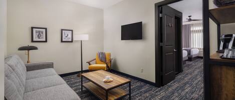A hotel room with a grey sofa, a wooden coffee table, a yellow armchair, a floor lamp, and a flat-screen TV on the wall.