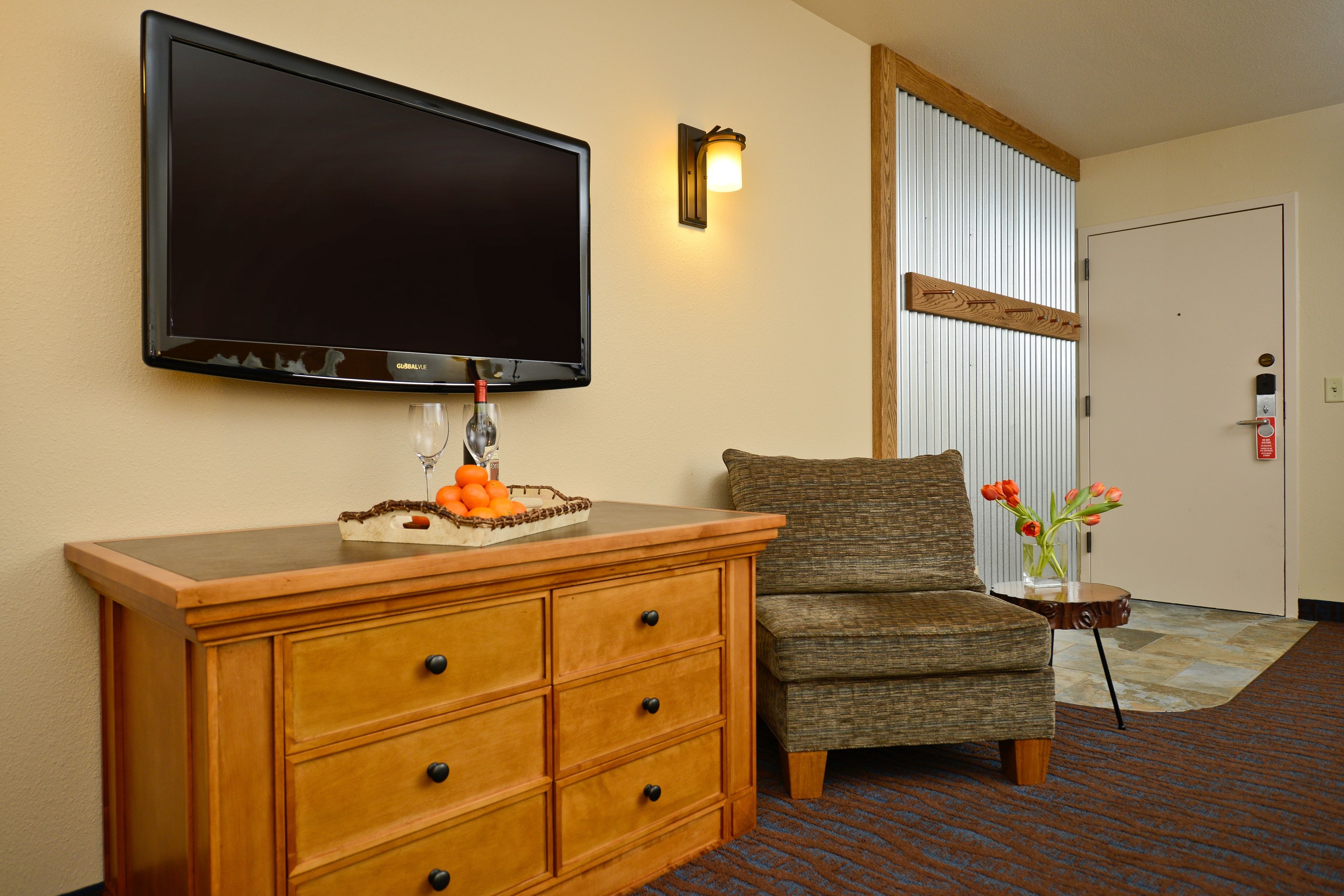 A hotel room with a flat-screen TV mounted on the wall, a wooden dresser with drawers, a gray armchair, a small side table with a vase of flowers, and a door with a wooden frame.