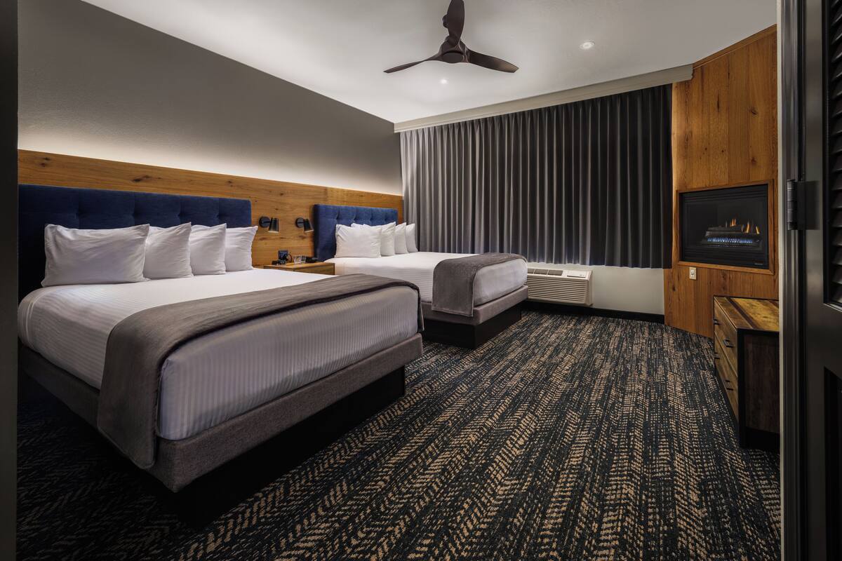 A hotel room with two beds, a ceiling fan, a fireplace, and a large window with curtains.