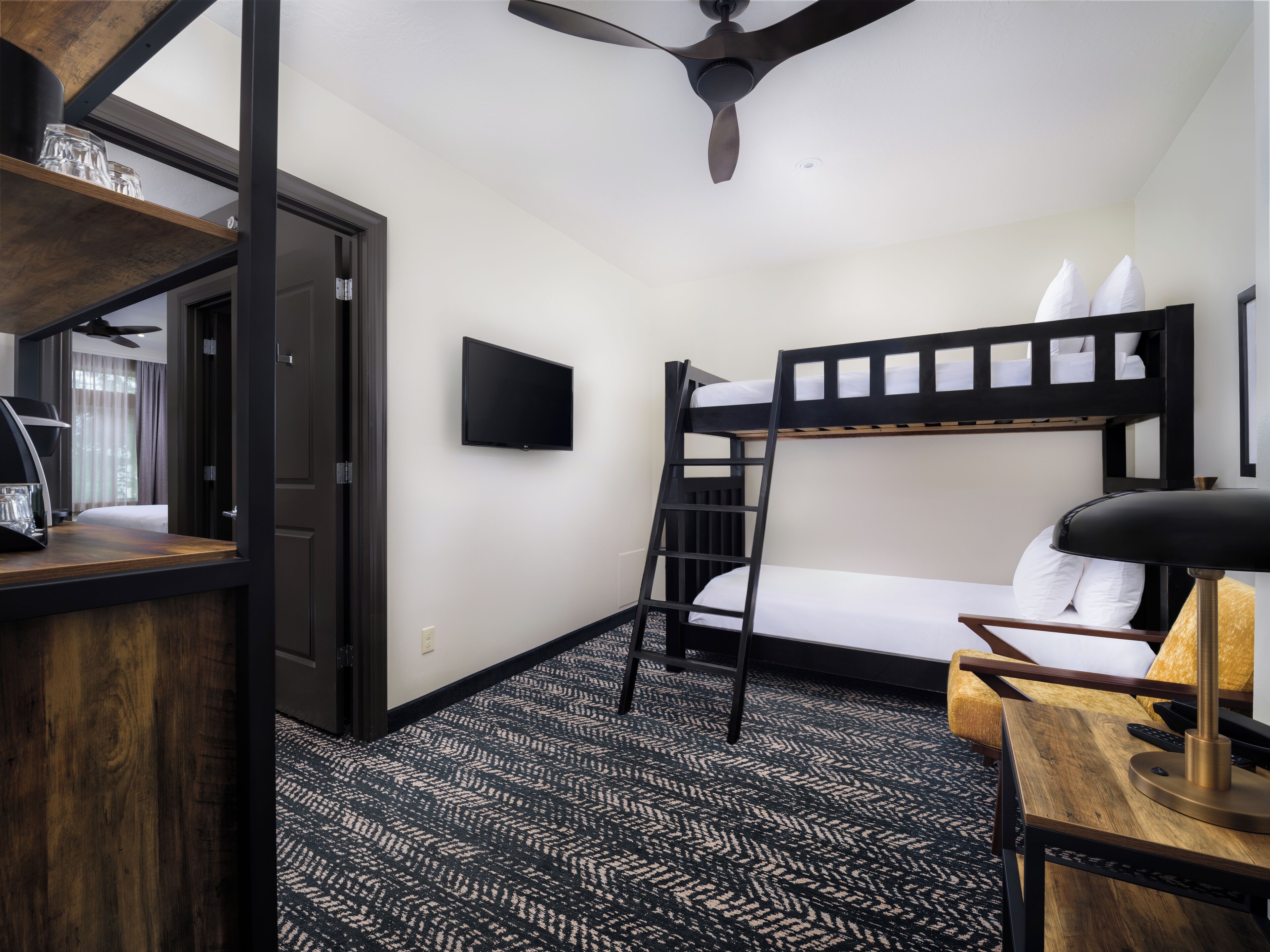 A compact hotel room with a bunk bed, a desk with a lamp, a television, and a ceiling fan.