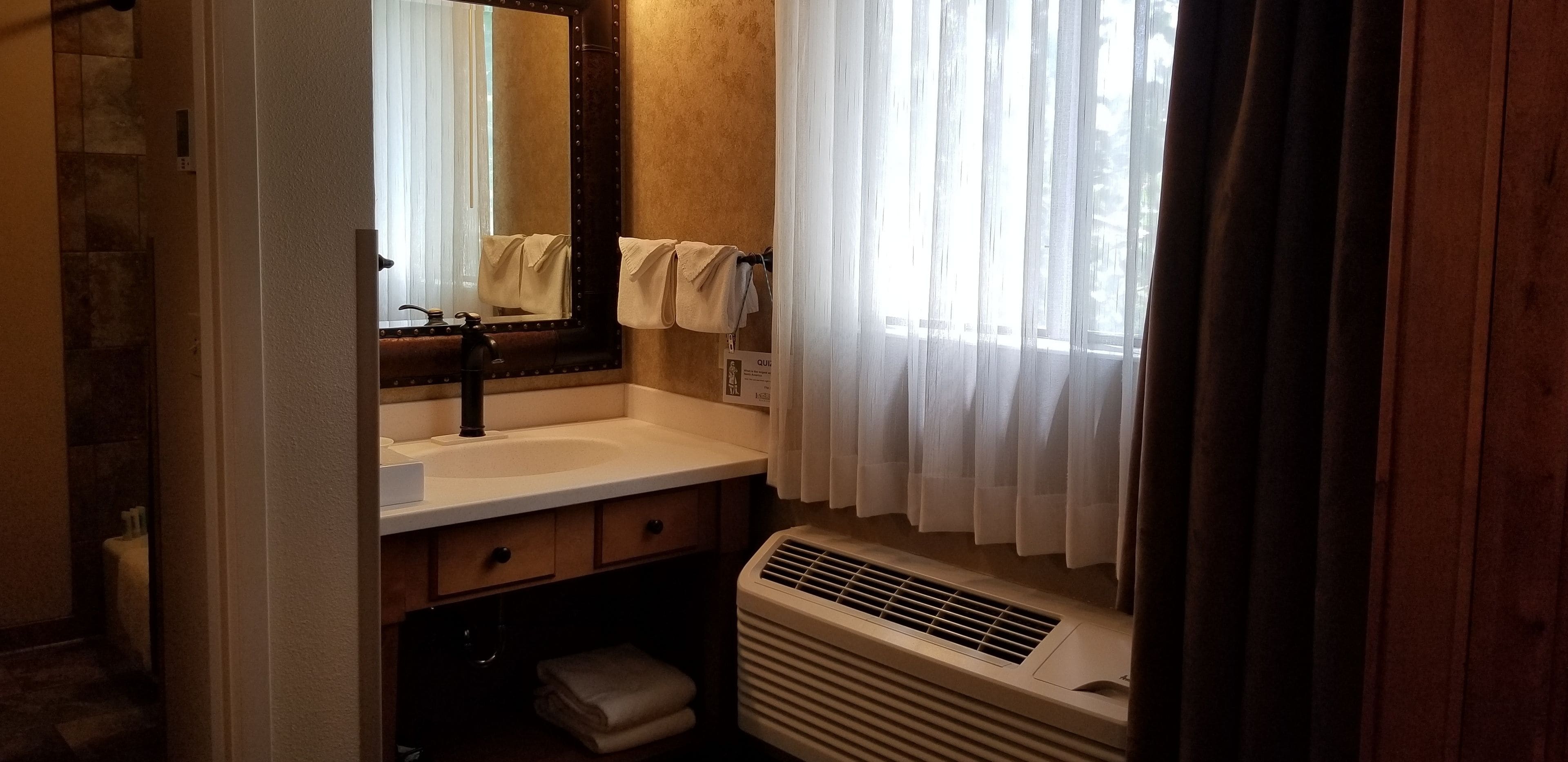 A bathroom with a white sink, a mirror with a dark frame, a towel rack with towels, a window with sheer curtains, and a wall-mounted heater.