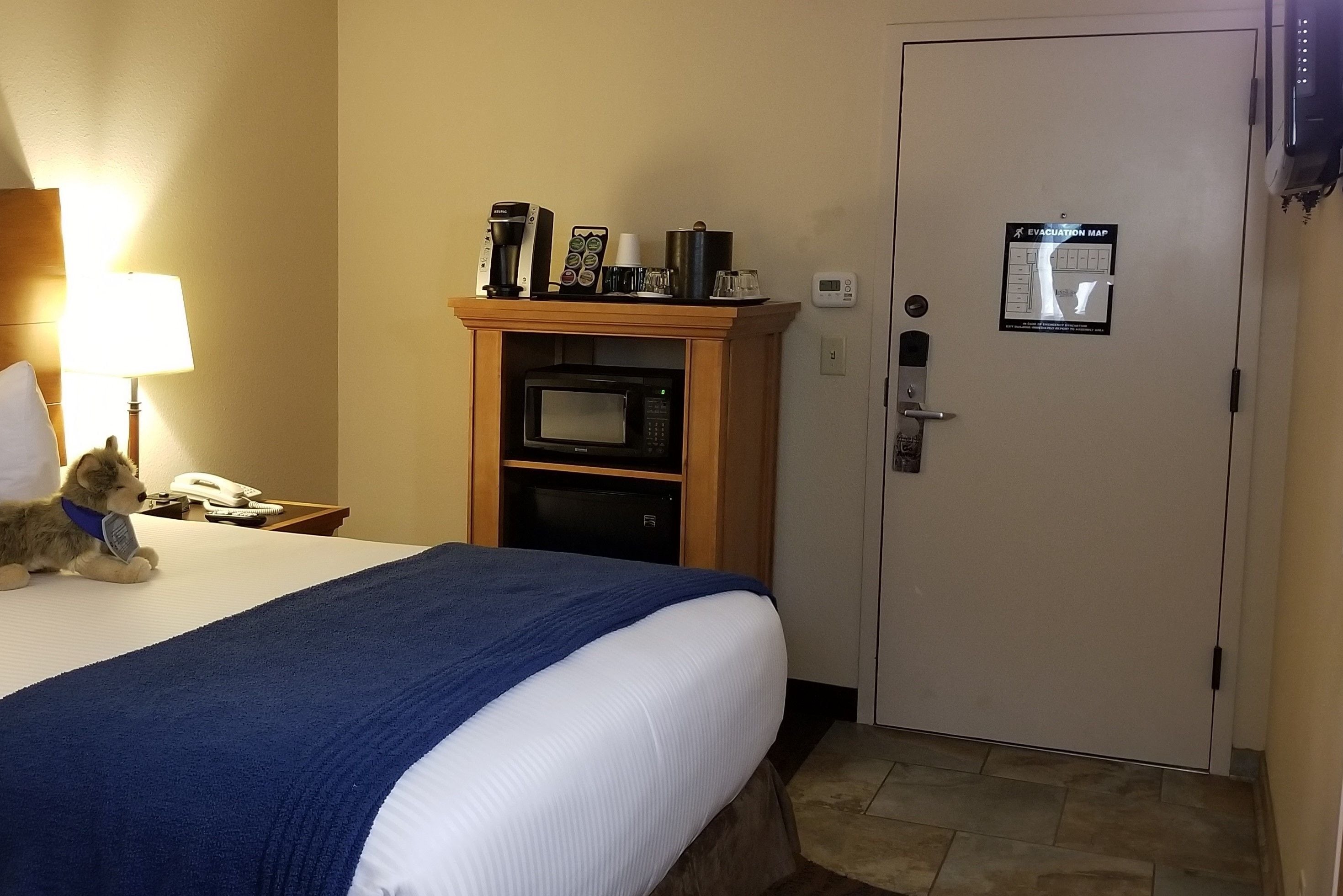 A hotel room with a bed, a microwave, a coffee maker, and a stuffed animal on the nightstand.