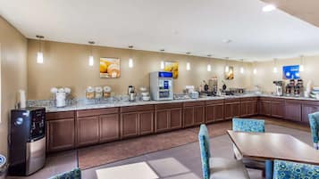 A hotel kitchen with a water dispenser, coffee maker, and a refrigerator. There are chairs and a table in the dining area.