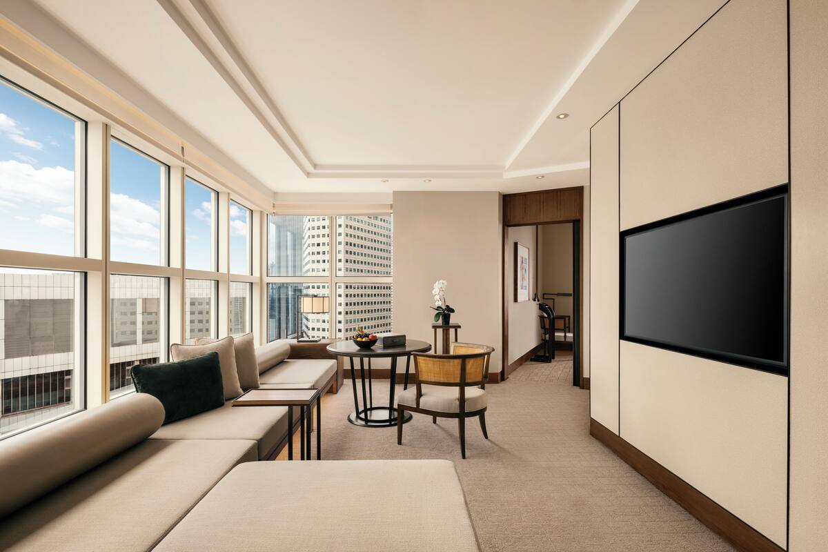 Una habitación de hotel moderna con un ventanal grande, un sofá, una mesita, una silla y un televisor de pantalla plana.