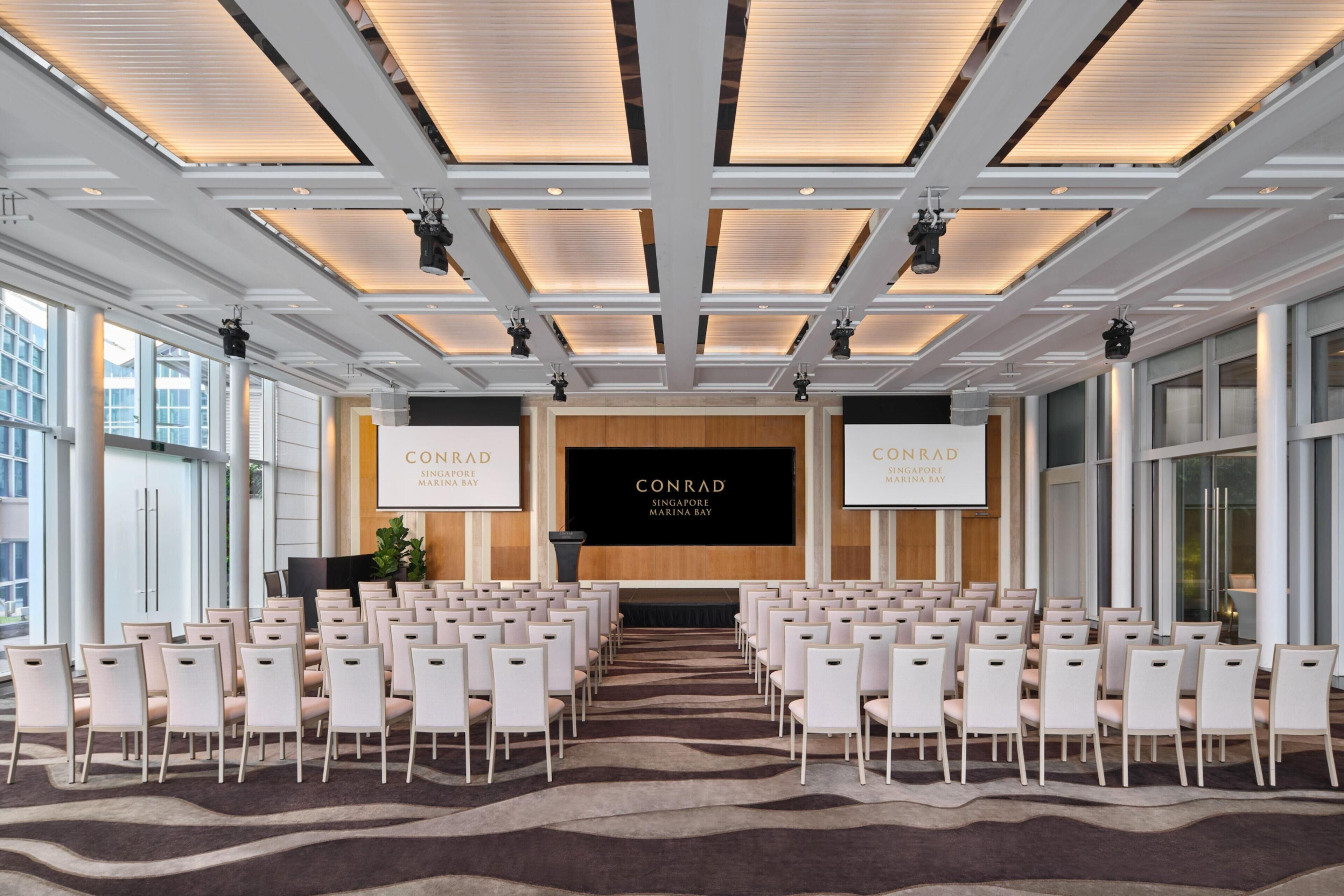 Une salle de conférence avec des rangées de chaises, un grand écran et une enseigne indiquant « CONRAD SINGAPORE MARINA BAY ».