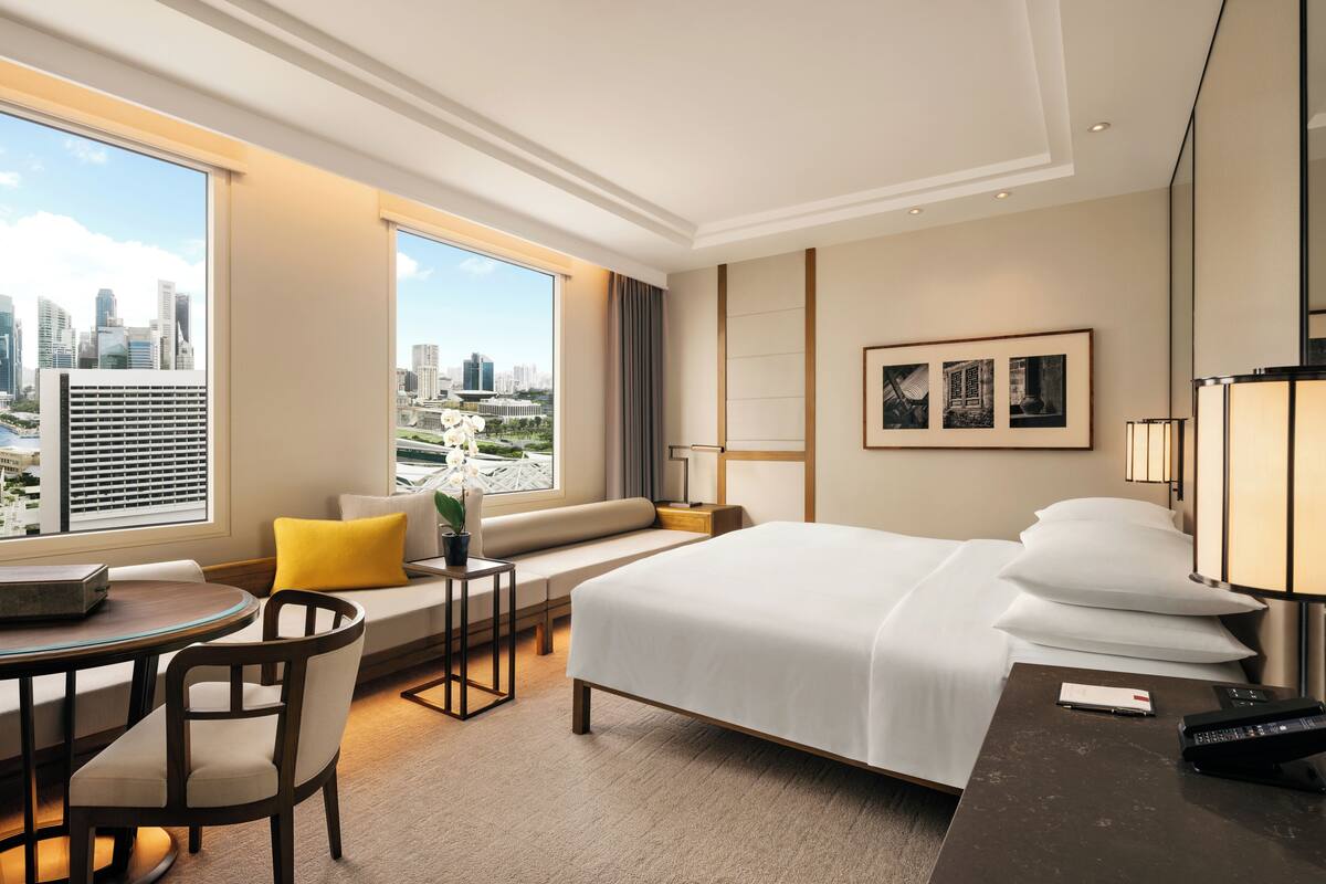 Habitación de hotel con una cama grande, zona de estar con sofá y sillones, una mesita y vistas a la ciudad a través de amplias ventanas.