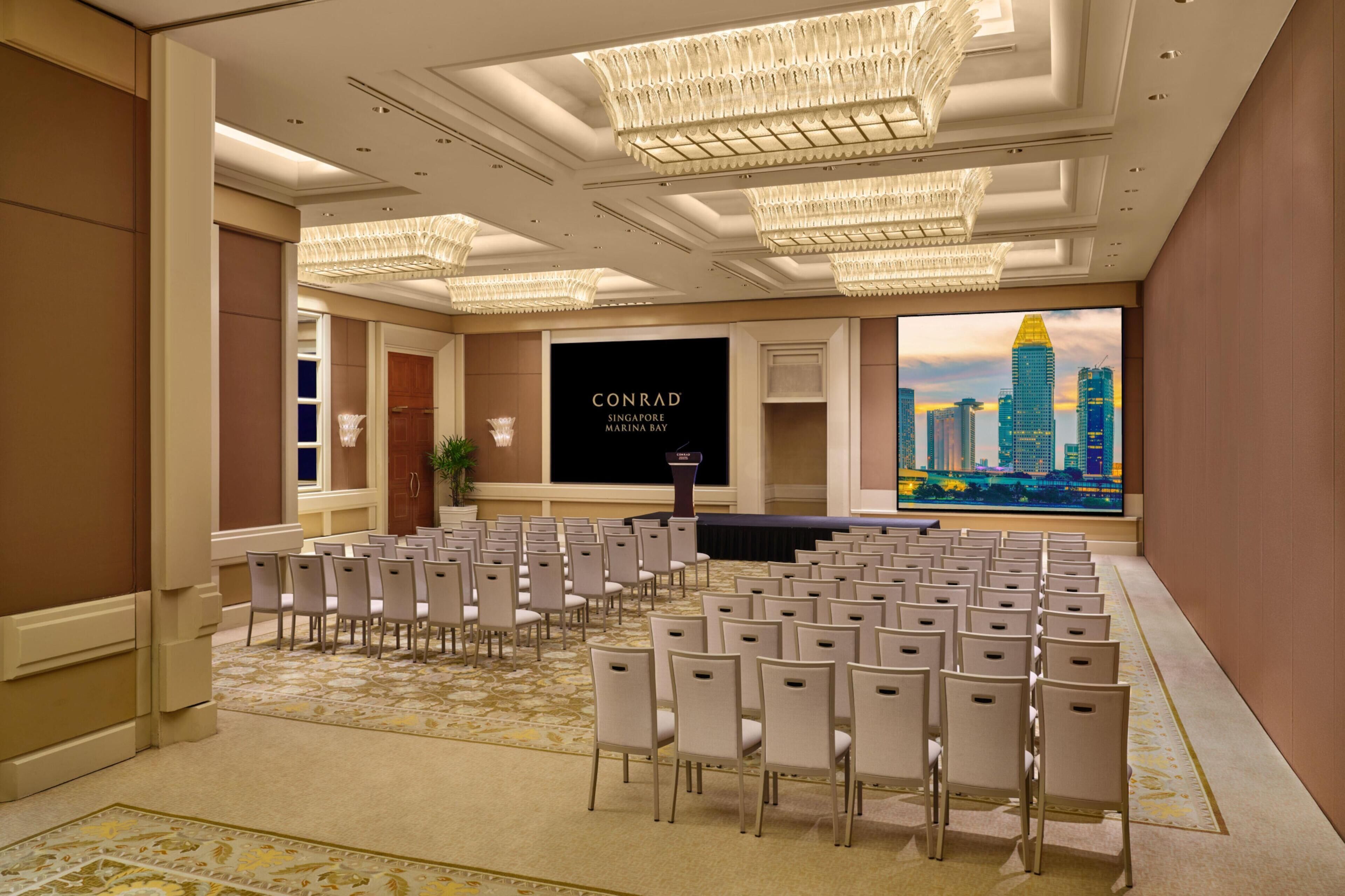 A conference room with rows of chairs, a podium, and a large screen displaying a cityscape.