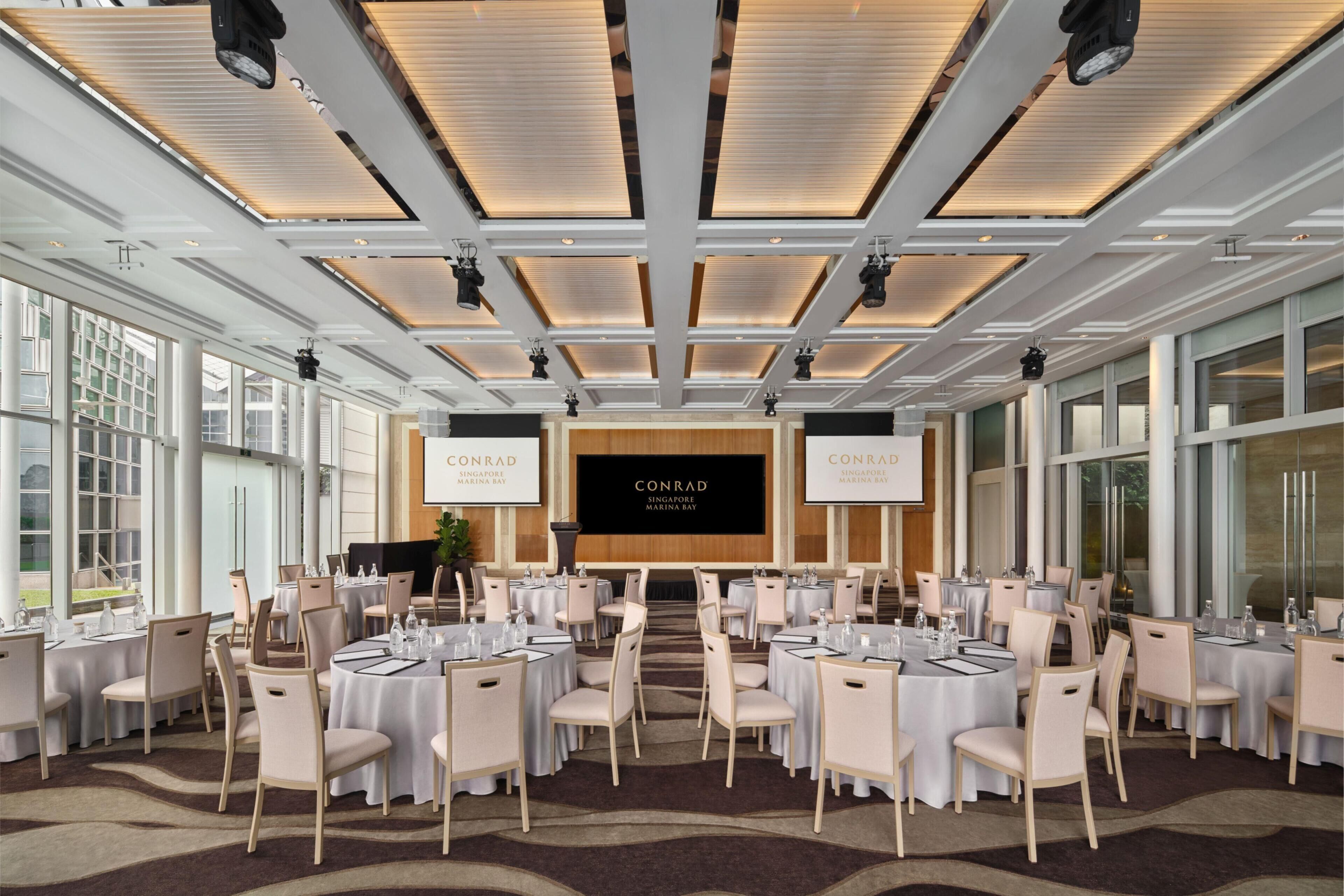 Une salle de conférence équipée de tables rondes, de chaises et d’un grand écran affichant le texte « CONRAD SINGAPORE MARINA BAY ».