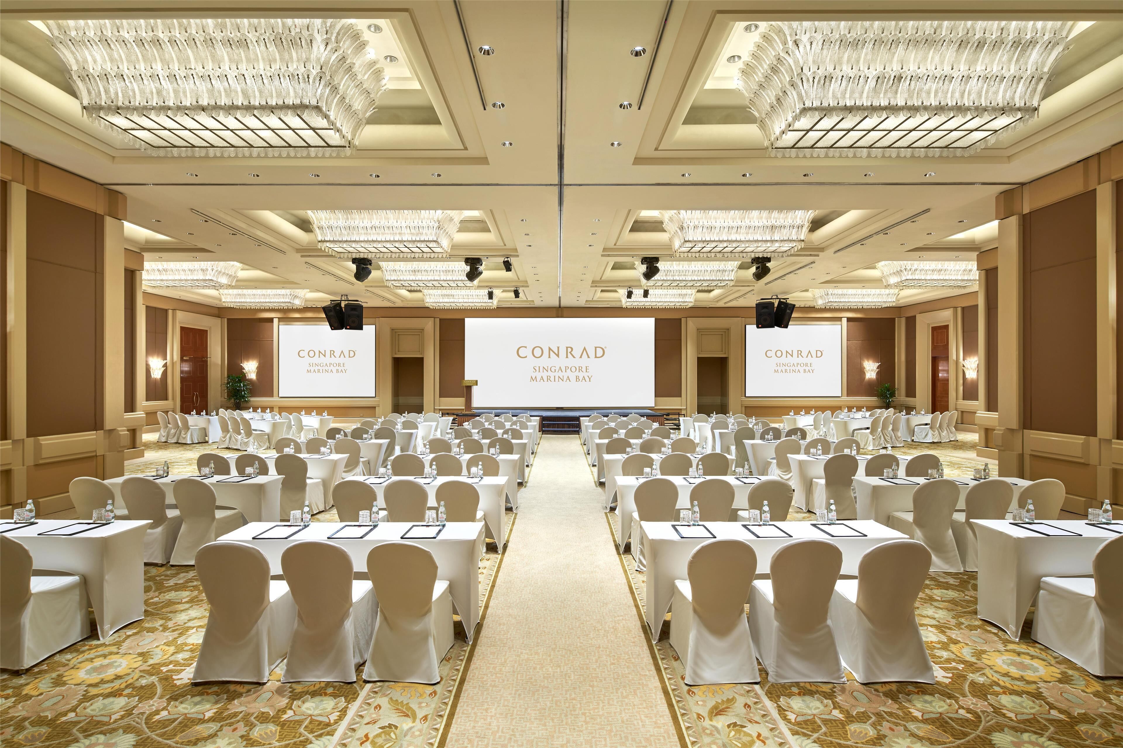 A large, well-lit conference room with rows of tables and chairs, a central stage with a screen displaying 'CONRAD SINGAPORE MARINA BAY', and a carpeted floor.