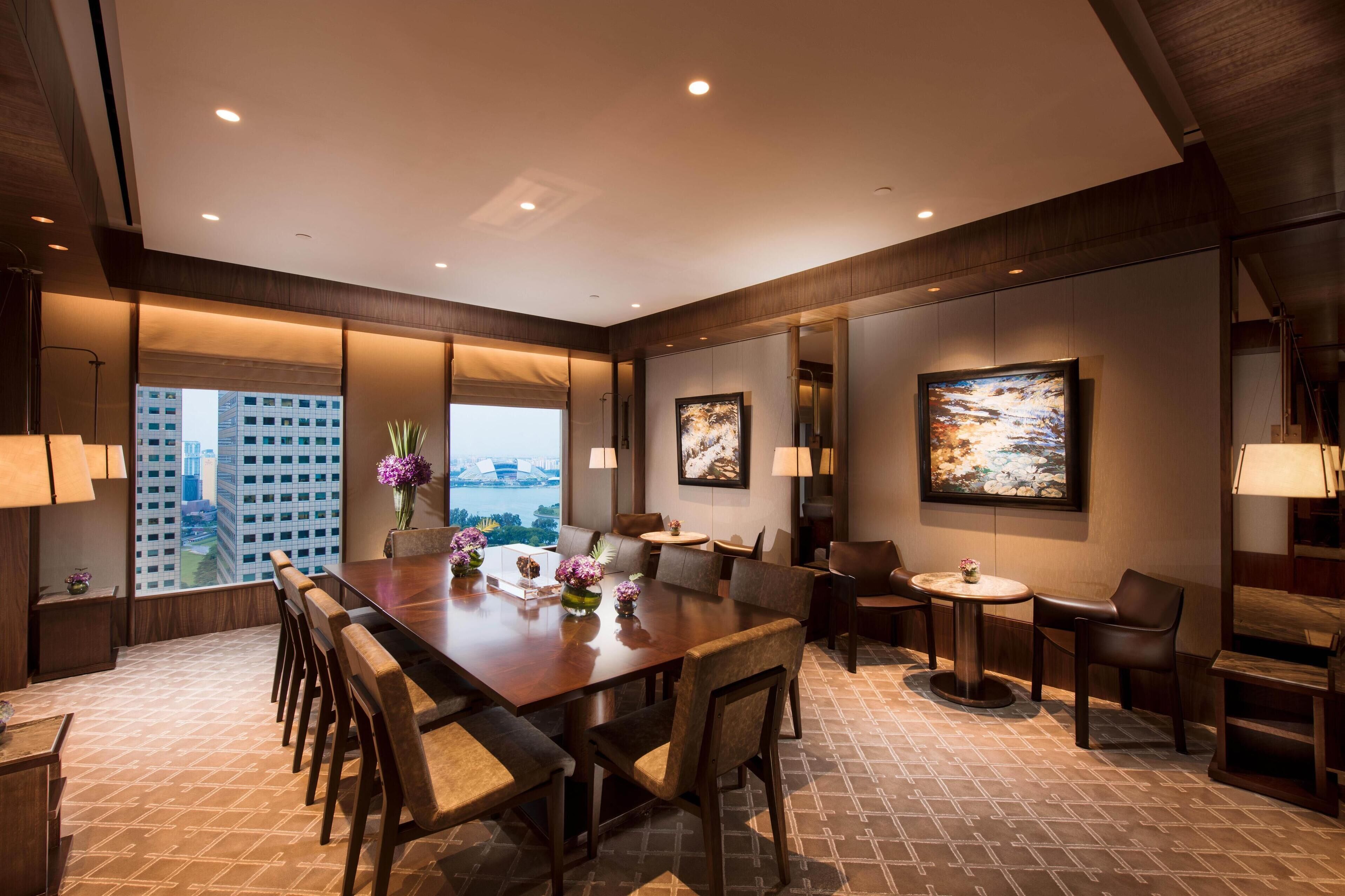 A modern hotel room with a large dining table, comfortable seating, artwork on the walls, and a view of a cityscape through large windows.