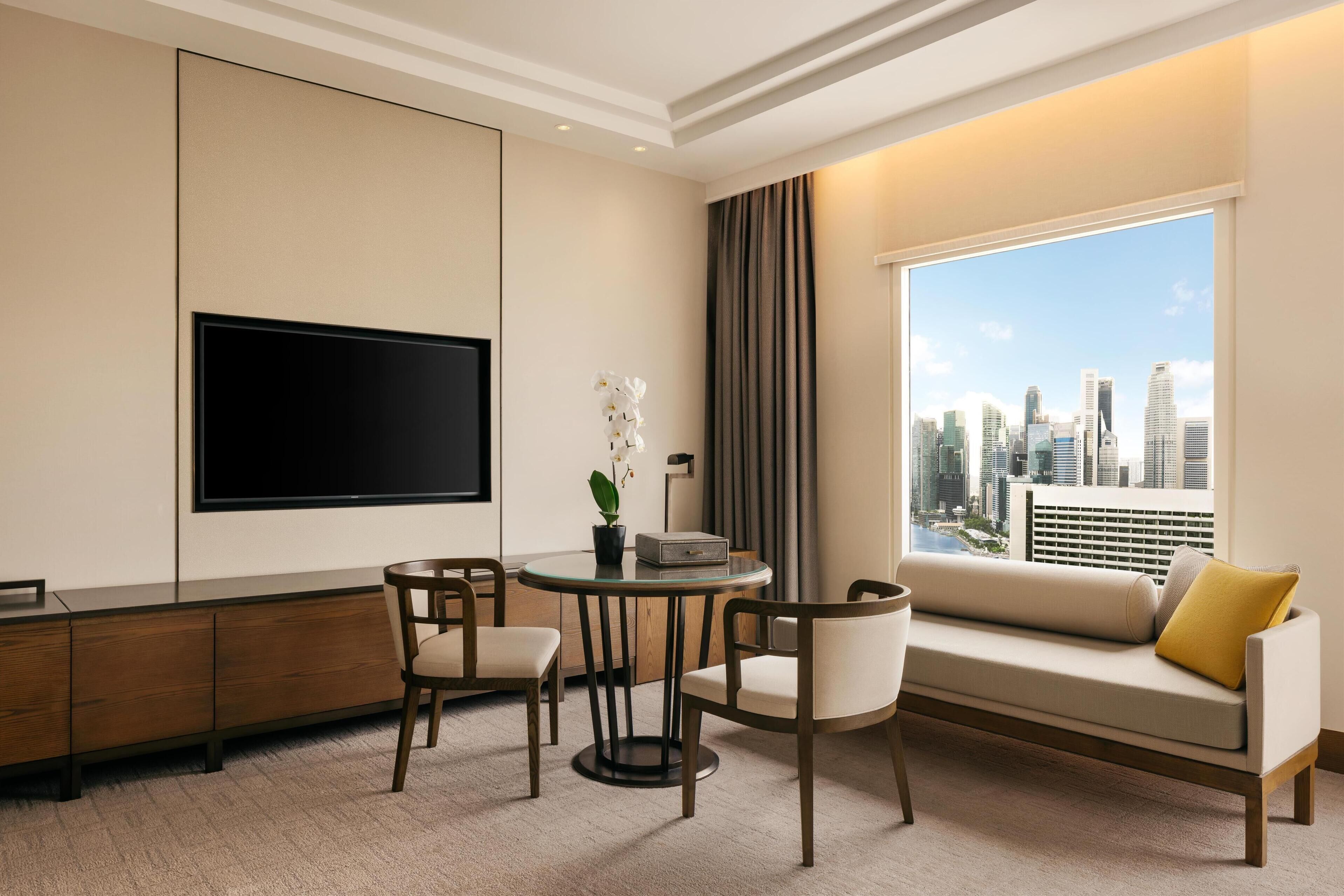Una camera d'albergo moderna con una grande televisione, un divano, un tavolo da pranzo con sedie e vista sulla città.