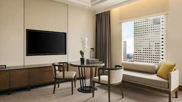 Una habitación de hotel moderna con una televisión de pantalla plana montada en la pared, una mesa redonda con sillas, un sofá con un cojín amarillo y un ventanal con vistas a la ciudad.