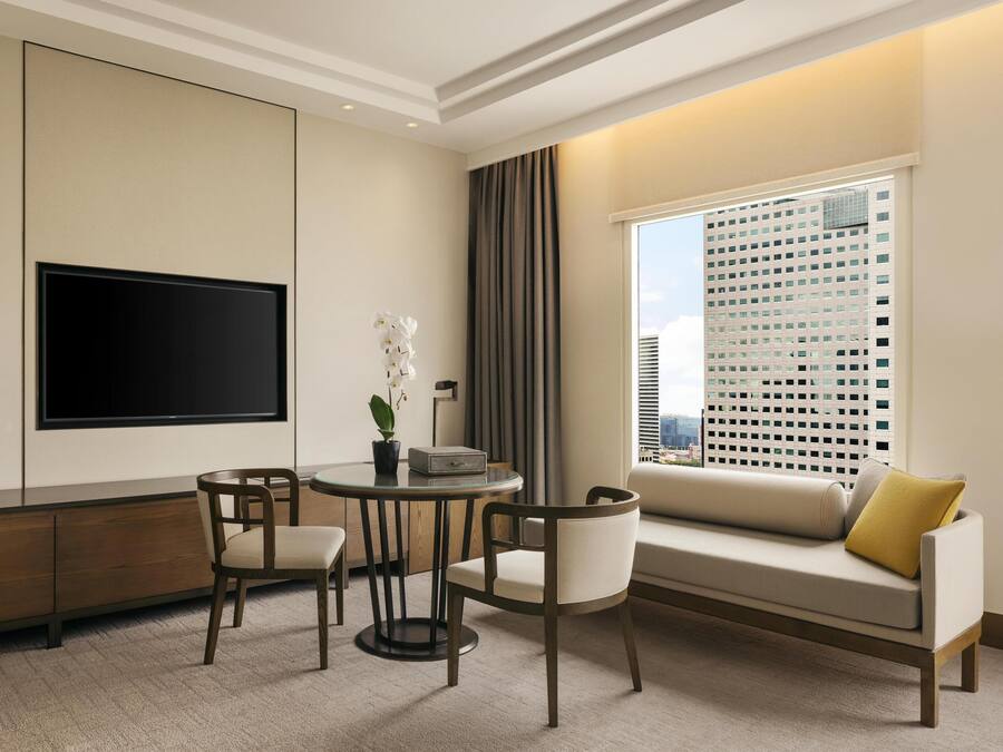 A modern hotel room with a flat-screen TV mounted on the wall, a round table with chairs, a sofa with a yellow pillow, and a large window with a city view.