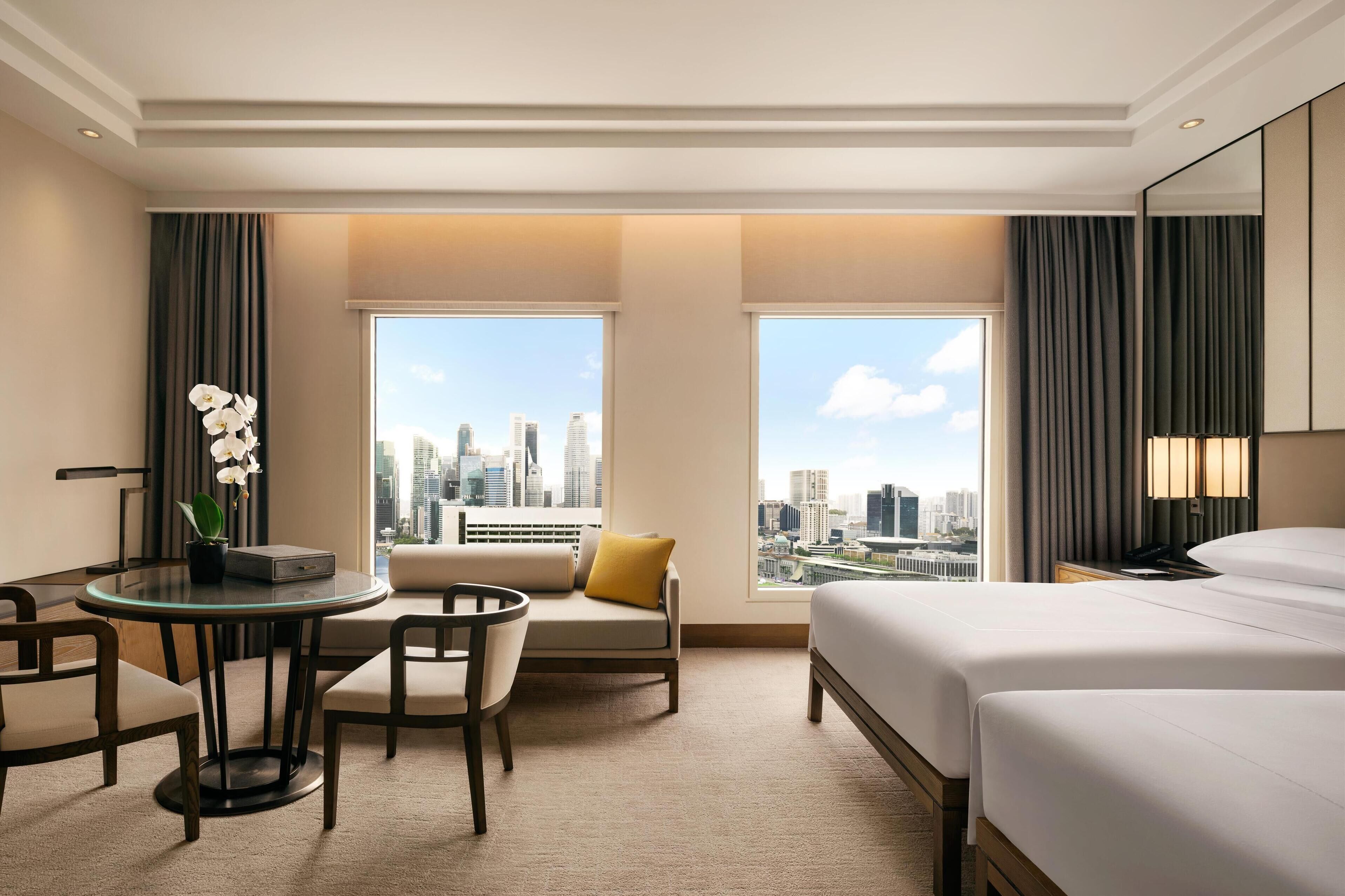 Habitación de hotel con vista a la ciudad, una mesa redonda de vidrio, dos camas y un área de descanso con un cojín amarillo.