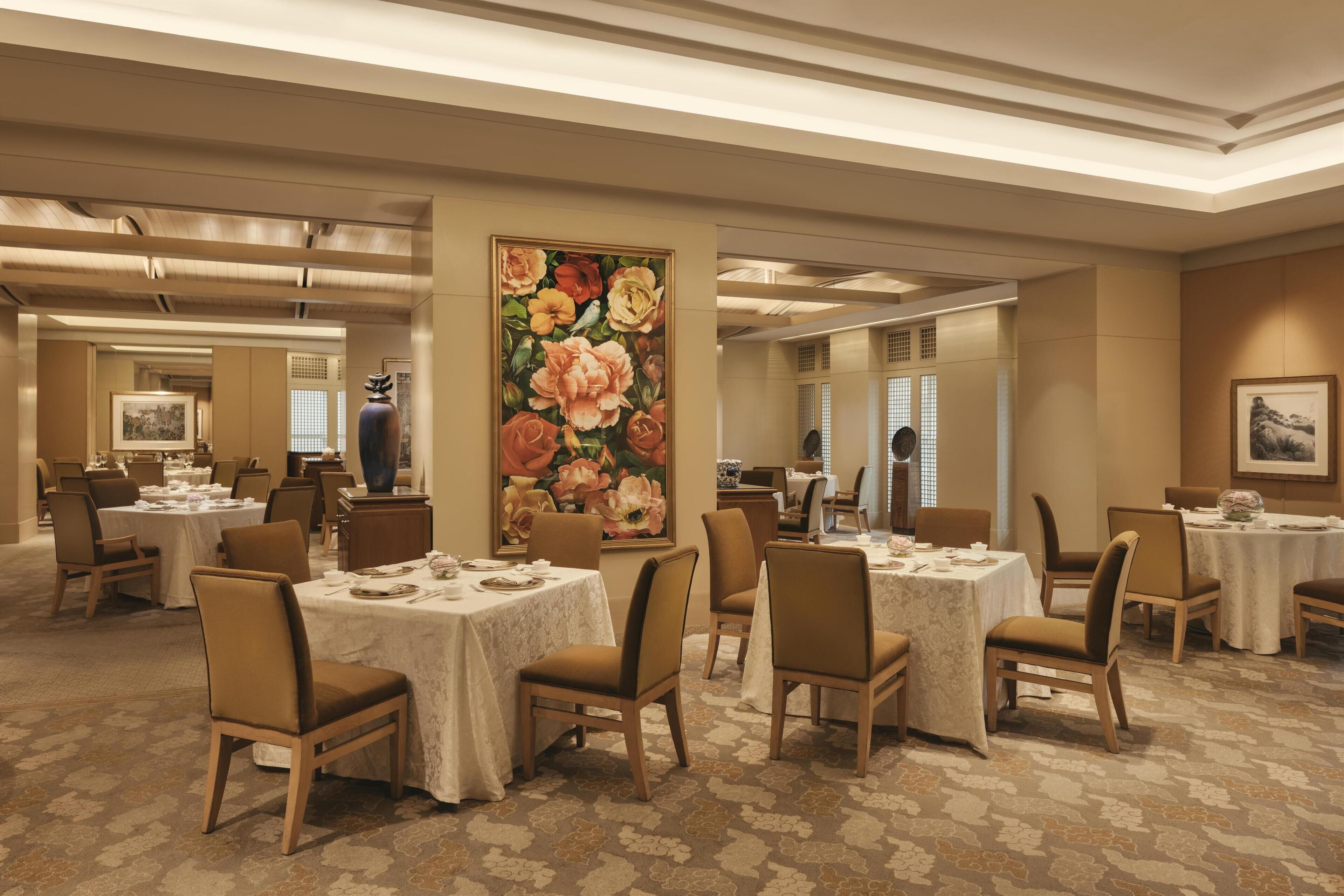 A hotel dining area with round tables, chairs, and a large floral painting on the wall.