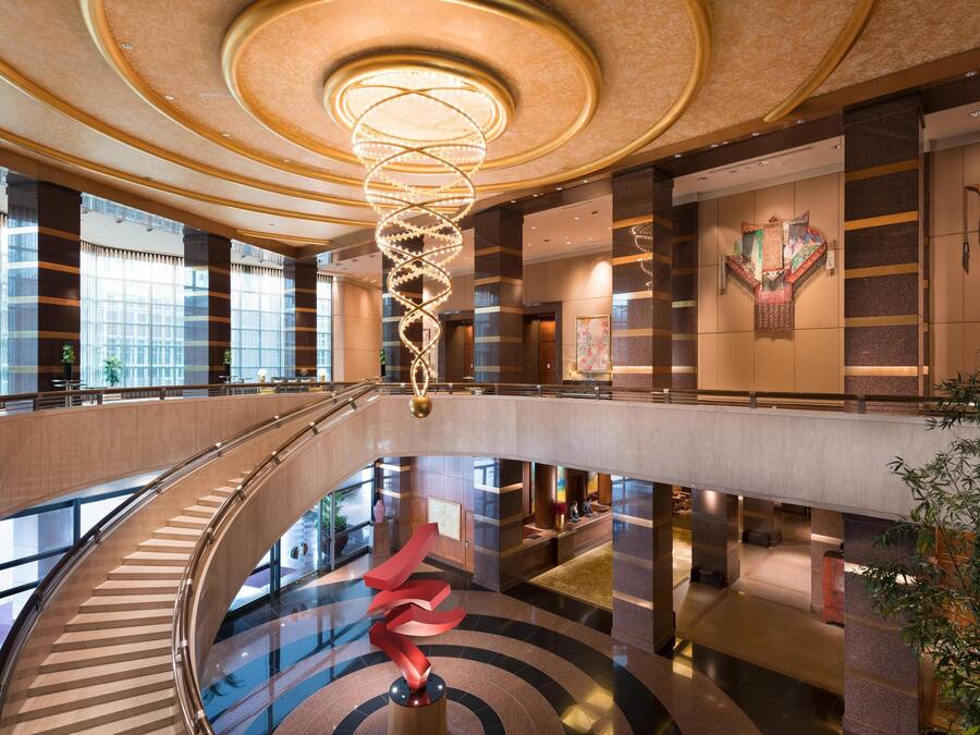 Un moderno vestíbulo de hotel con una lámpara espiral central, una escalera de cristal y una recepción.